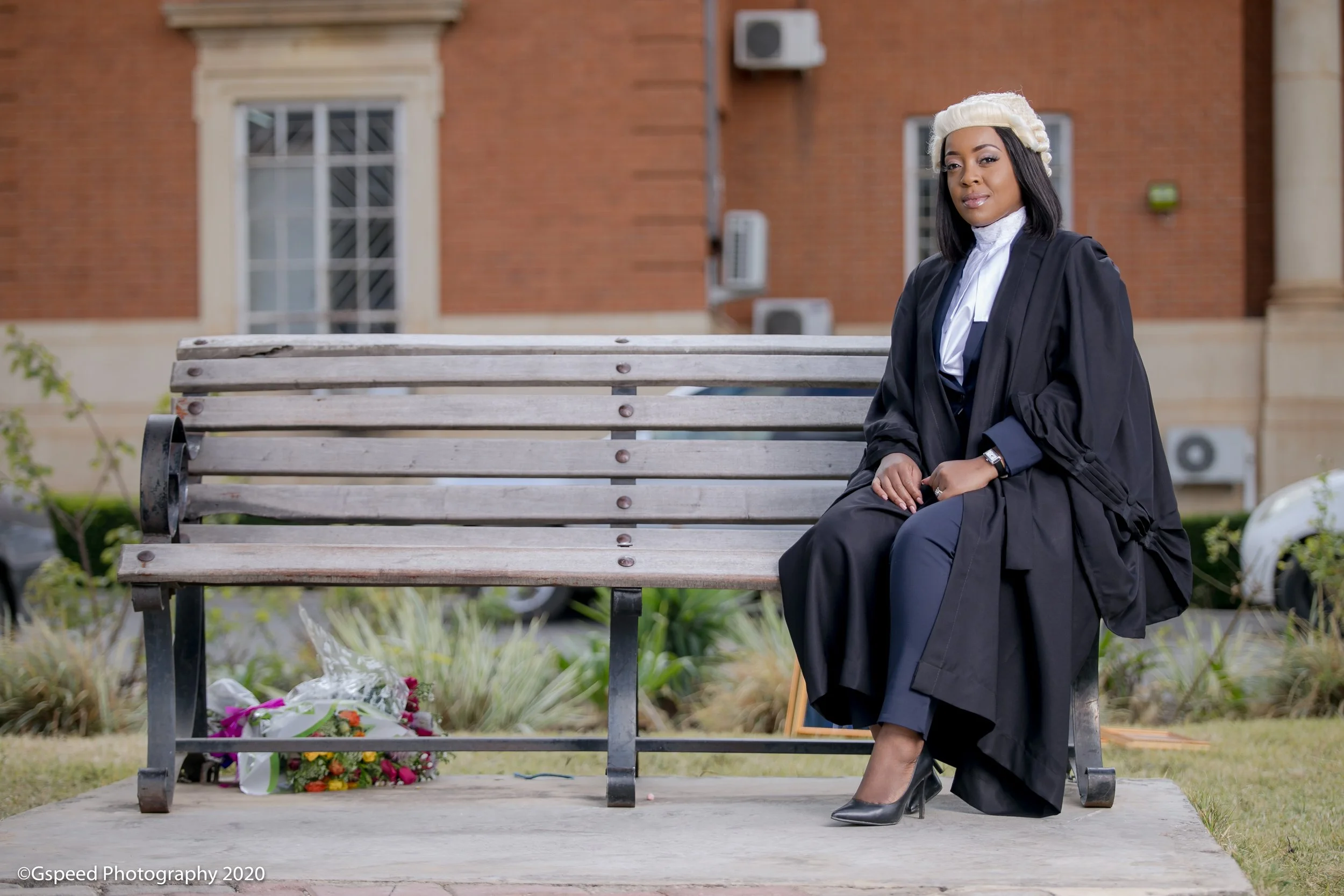 A woman in graduation cap and gown sitting on a park bench with flowers laid at the base of the bench, in front of a brick building.