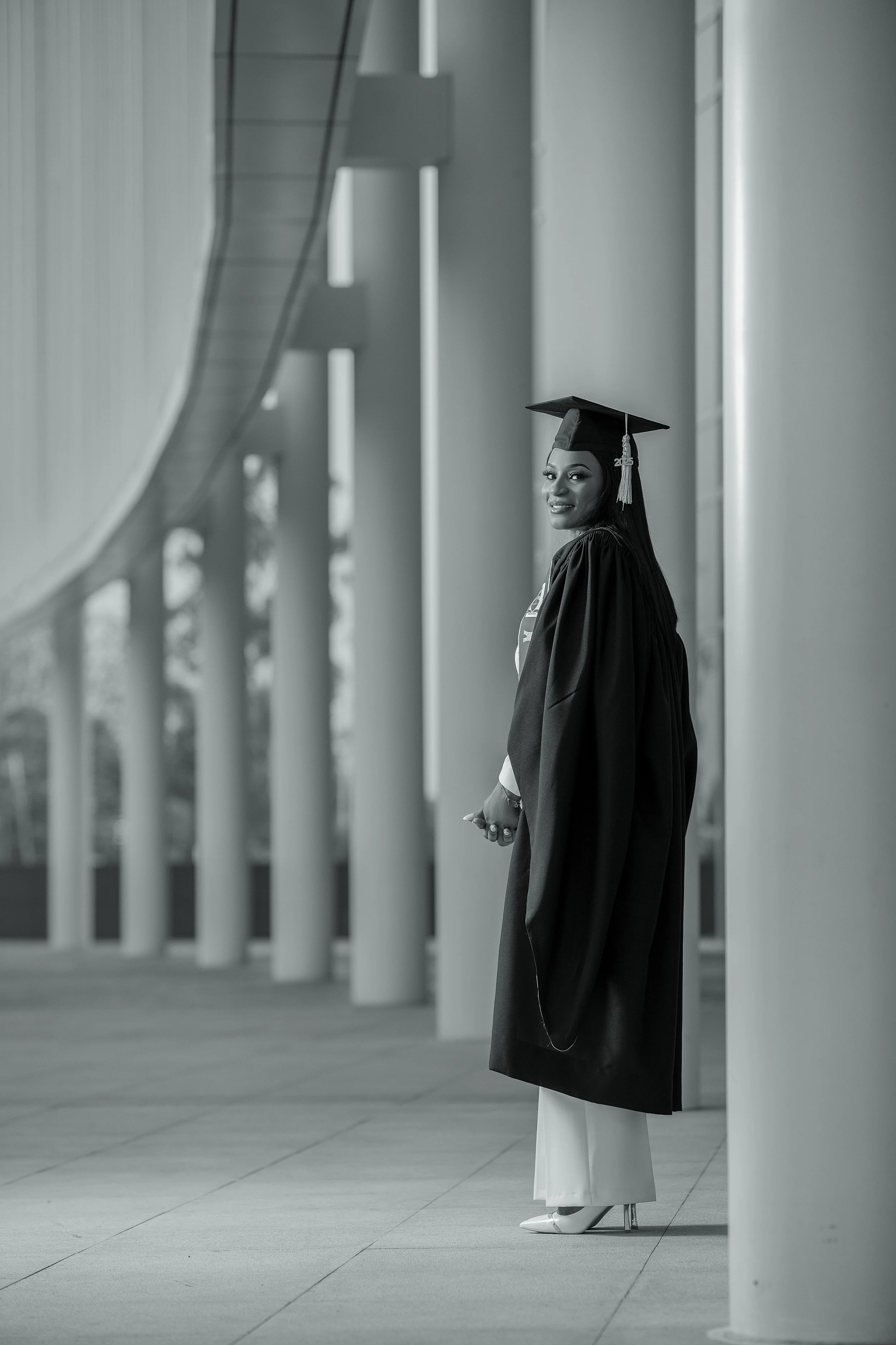 A woman in a graduation cap and gown, smiling and standing outside a modern building with tall, narrow windows and vertical architectural details.