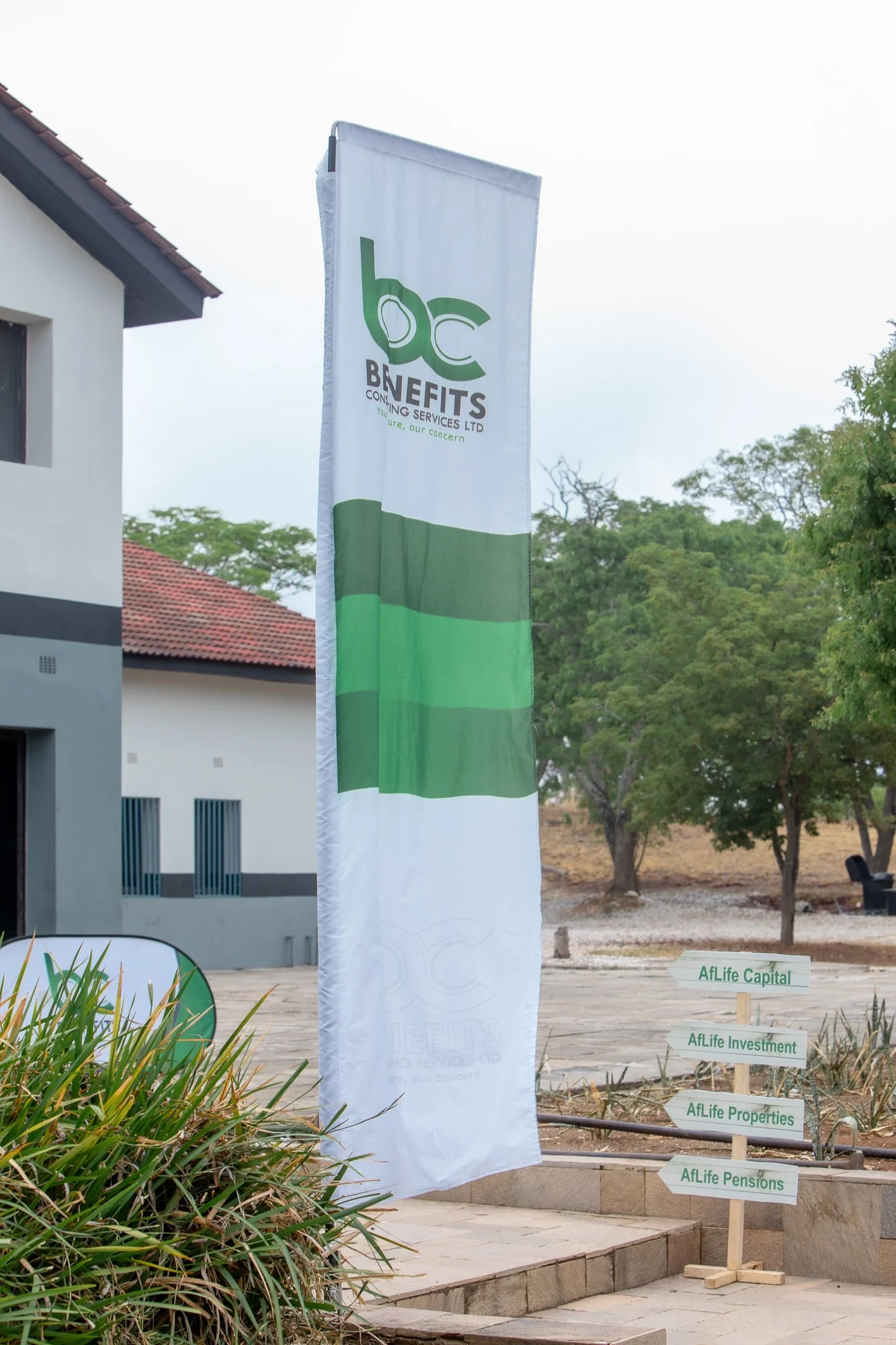 Outdoor scene featuring a tall white banner with green and black text that reads 'bc BENEFITS CONTINUING SERVICES LTD' with the slogan 'You are, our concern' beneath it. In the background, trees and a building with a tiled roof, and outdoor signage d