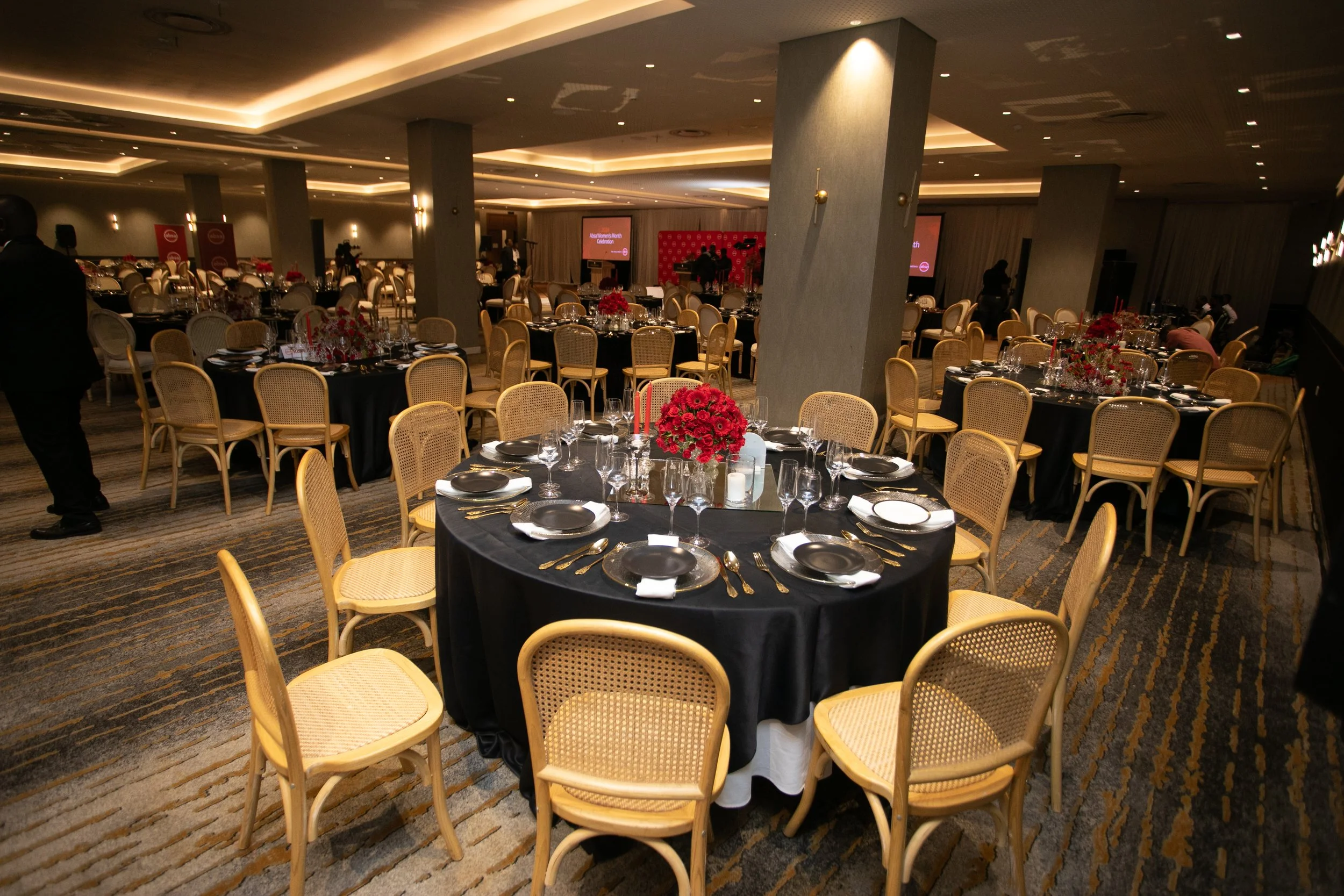 Elegant banquet hall set up for a celebration with round tables covered in black tablecloths, decorated with red floral centerpieces, and arranged with glassware, cutlery, and black plates. The room features warm lighting, beige chairs with woven bac