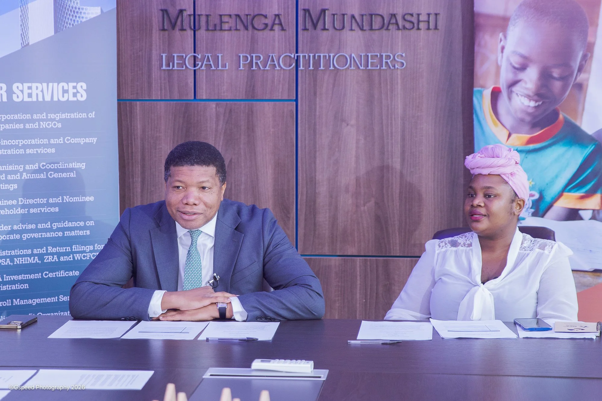 Two people sitting at a conference table in front of a wood panel wall with signs that read 'Mulenga Mundashi Legal Practitioners' and a large photo of a smiling woman in colorful attire. Documents and a pen are on the table.