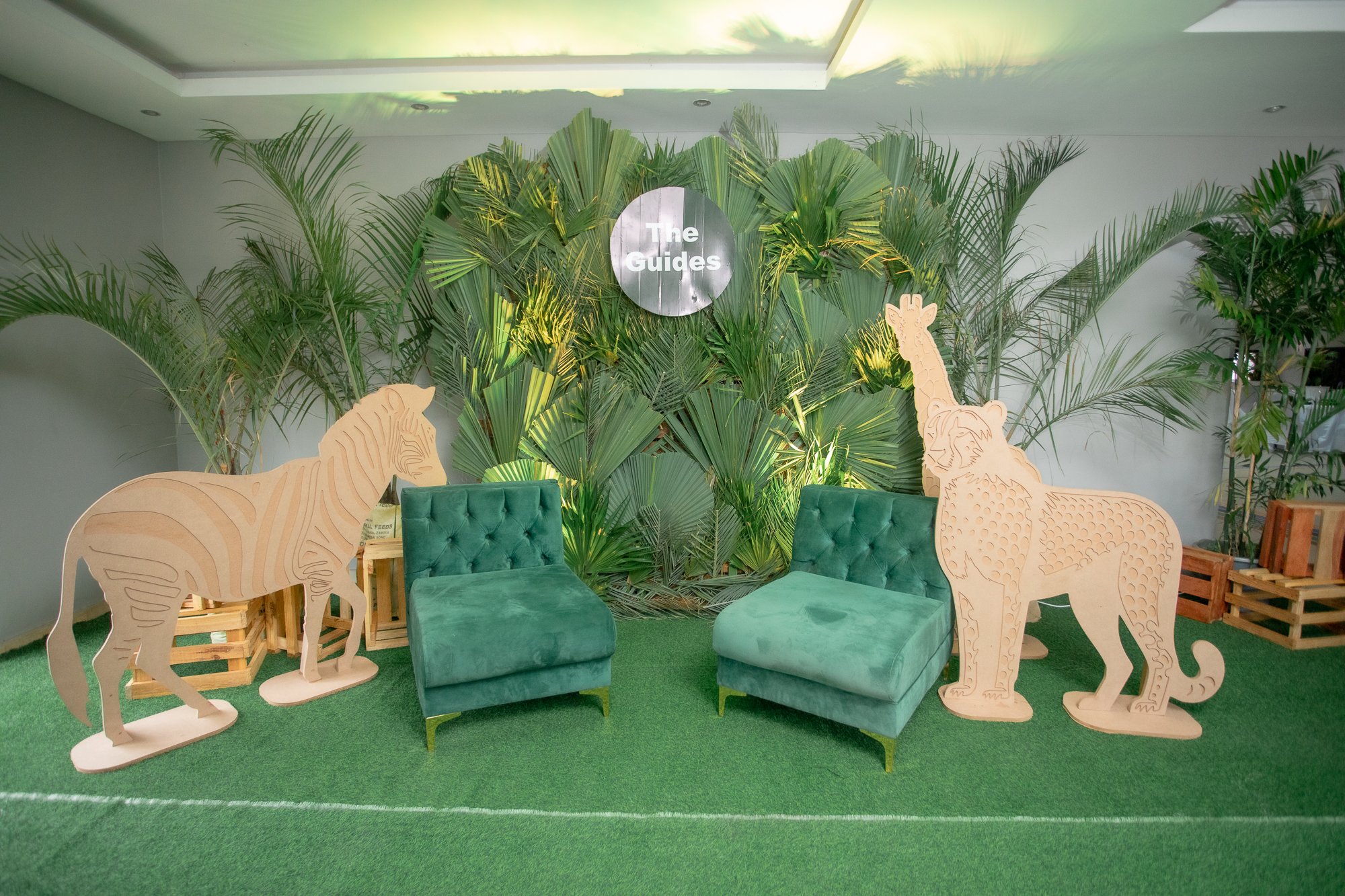 Indoor space decorated with large green tropical plants and cardboard cutouts of a zebra and a lion, with a circular sign in the center reading 'The Guides'. There are green cushioned benches and a green carpeted floor.