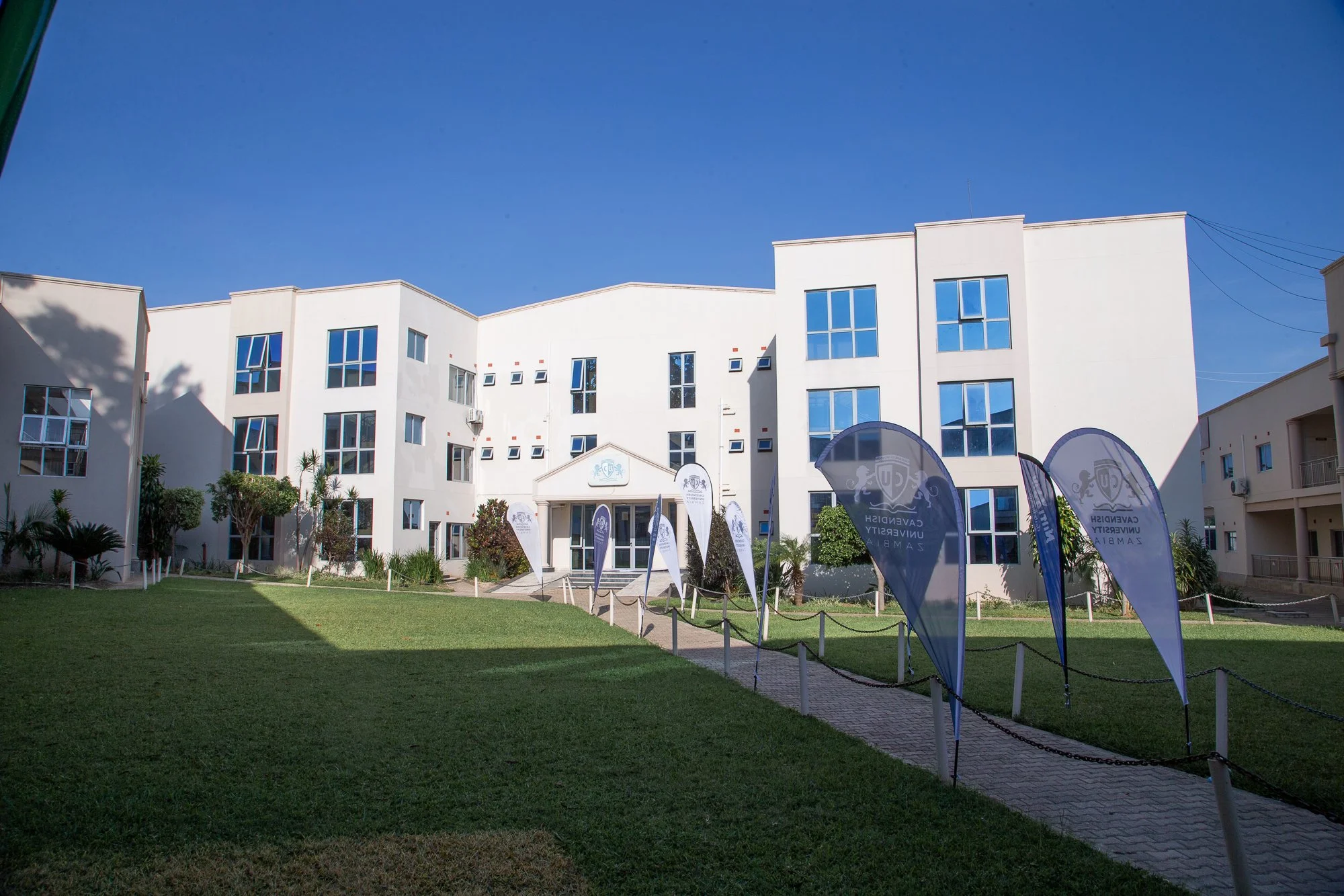 A white multi-story building with large windows under a clear blue sky, with a green lawn and flags at the entrance.