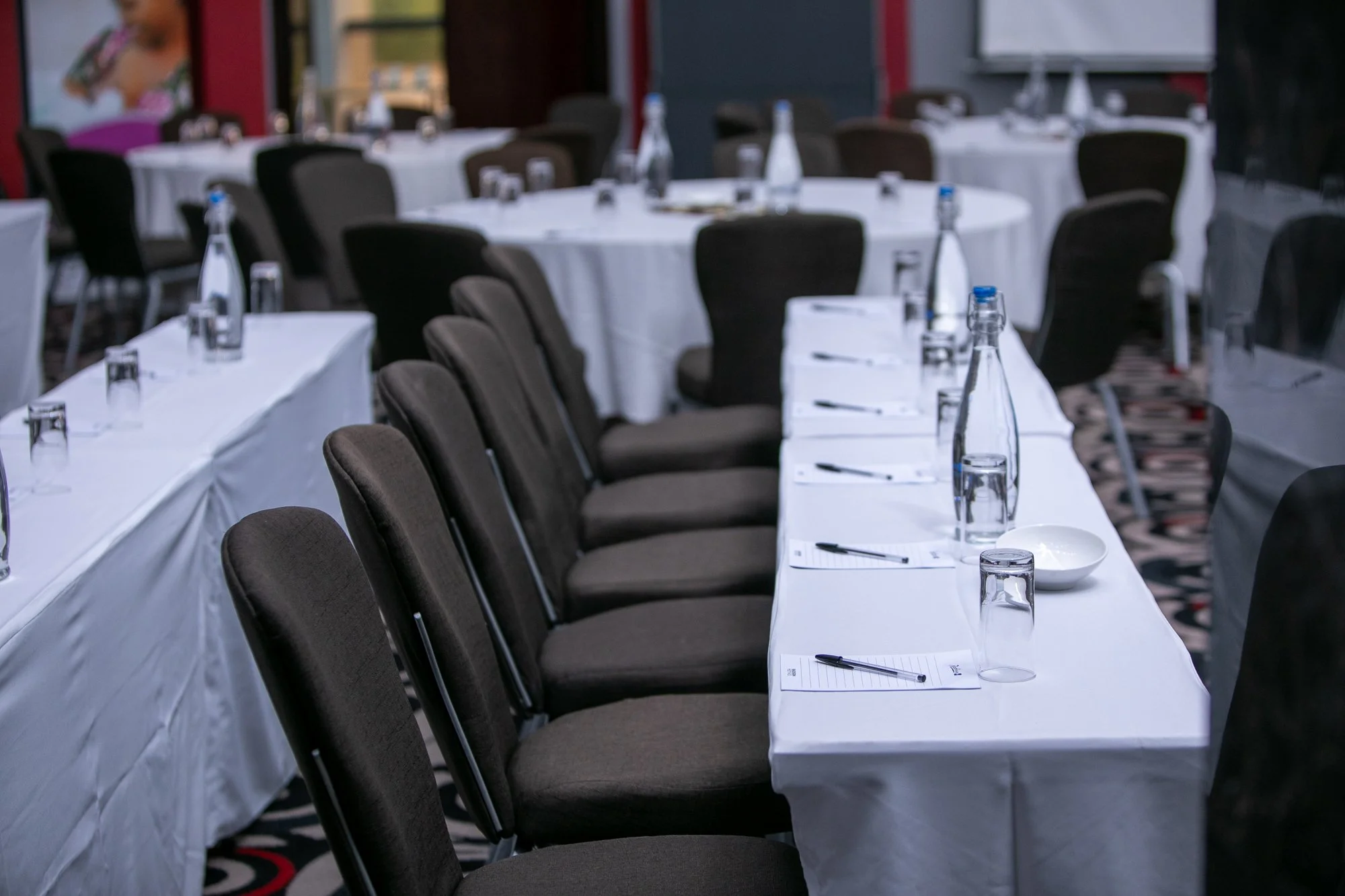 Conference room with round tables and black chairs set up for a meeting. Each table has water bottles, glasses, notepads, and pens, with some tables covered with white tablecloths.