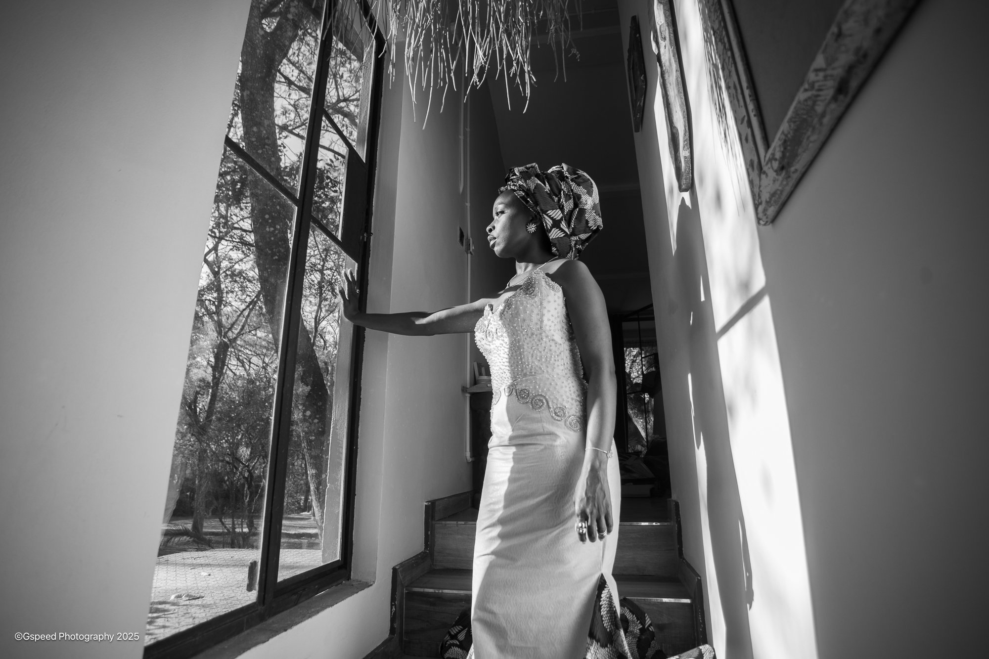 A woman wearing a patterned headwrap and sleeveless top, standing indoors and looking out a window with trees outside, with sunlight casting shadows inside.