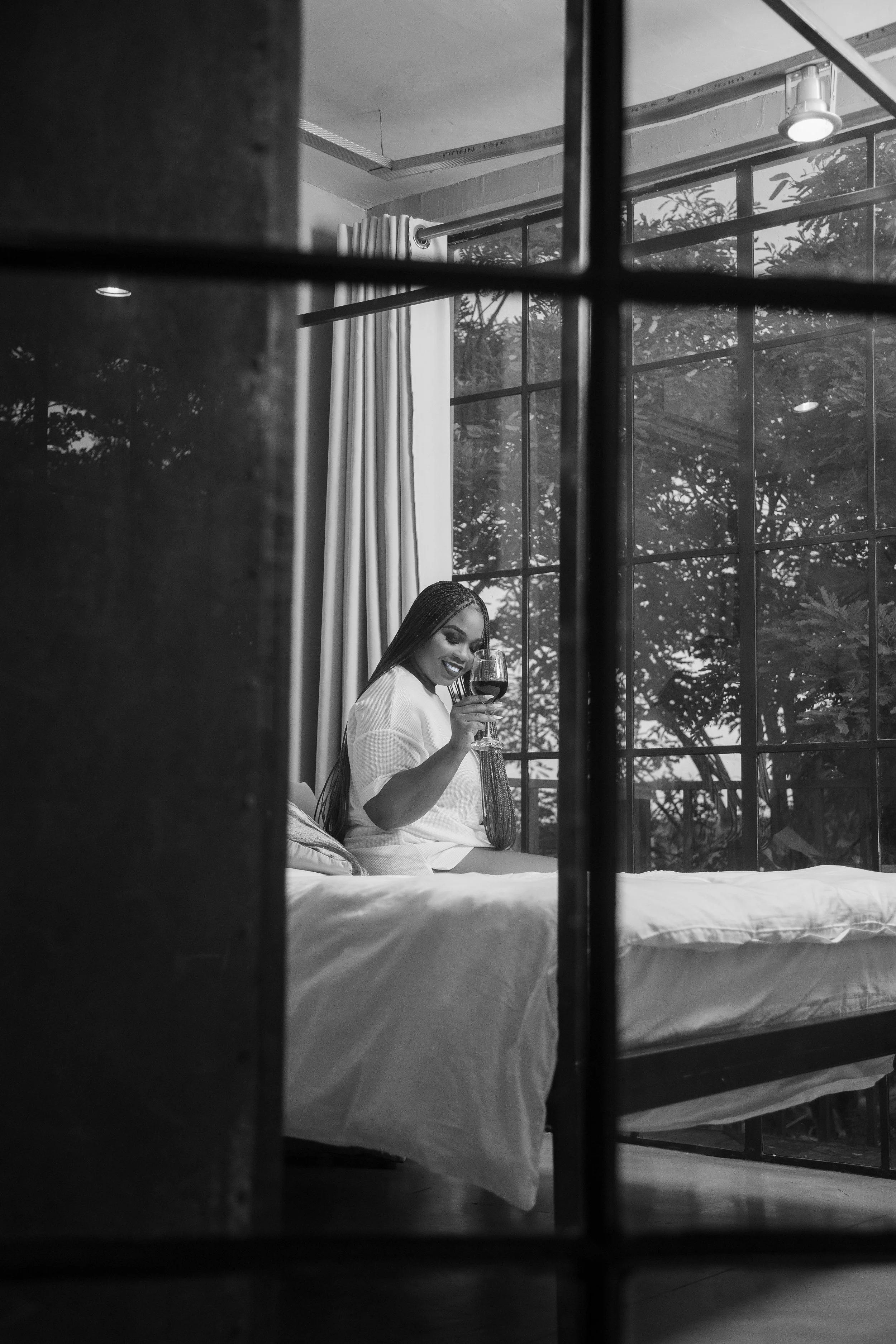 A woman sitting on a bed, smiling and holding a glass of red wine, viewed through the bars of a metal bed frame in a bedroom with large windows and curtains.