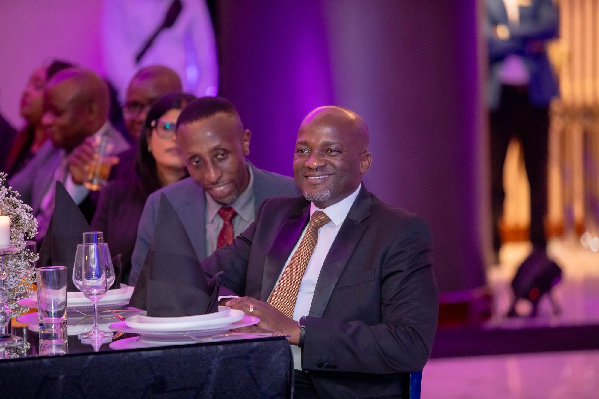 Group of people in formal attire sitting at a table during an event, with purple and pink lighting.