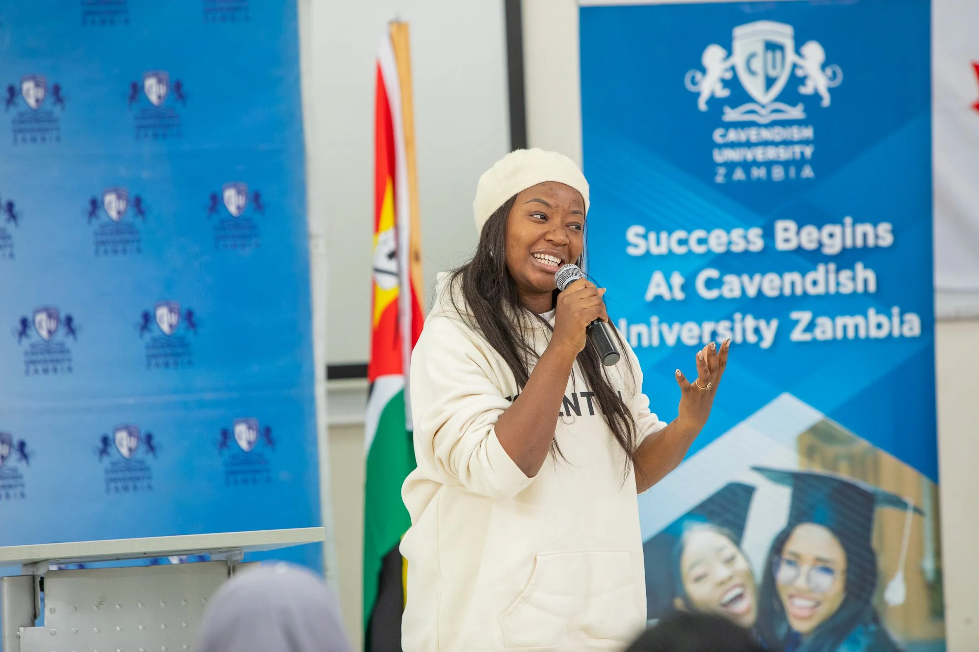 Young woman with a beige beret speaking into a microphone at an event, standing in front of blue banners with the Cavendish University Zambia logo and the slogan "Success Begins at Cavendish University Zambia."