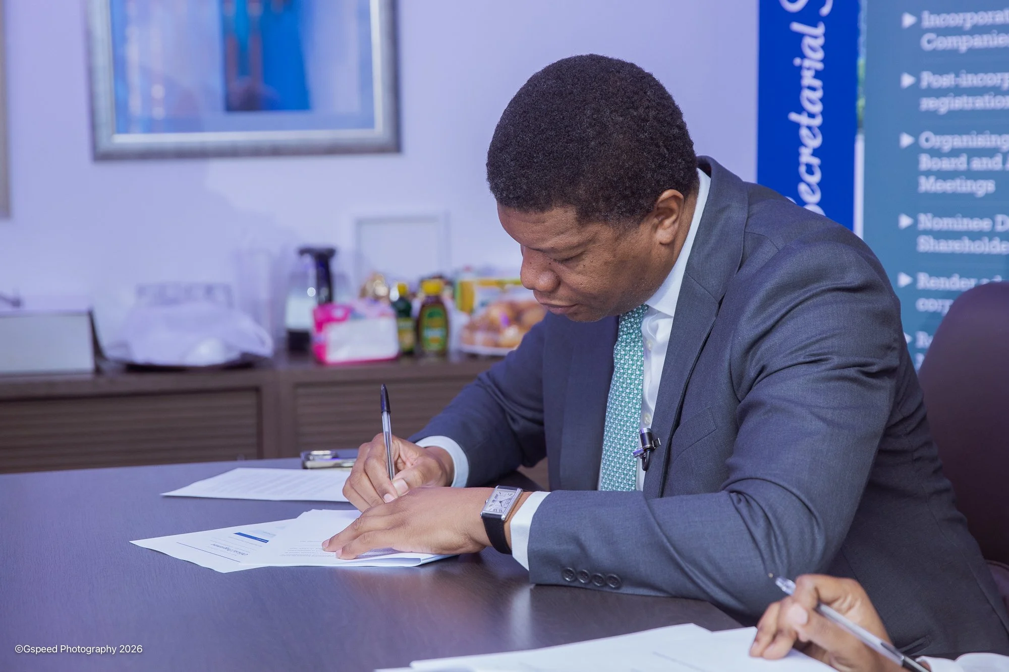 A man in a suit seated at a desk, signing a document, with papers and a pen in his hand, in a professional office setting.