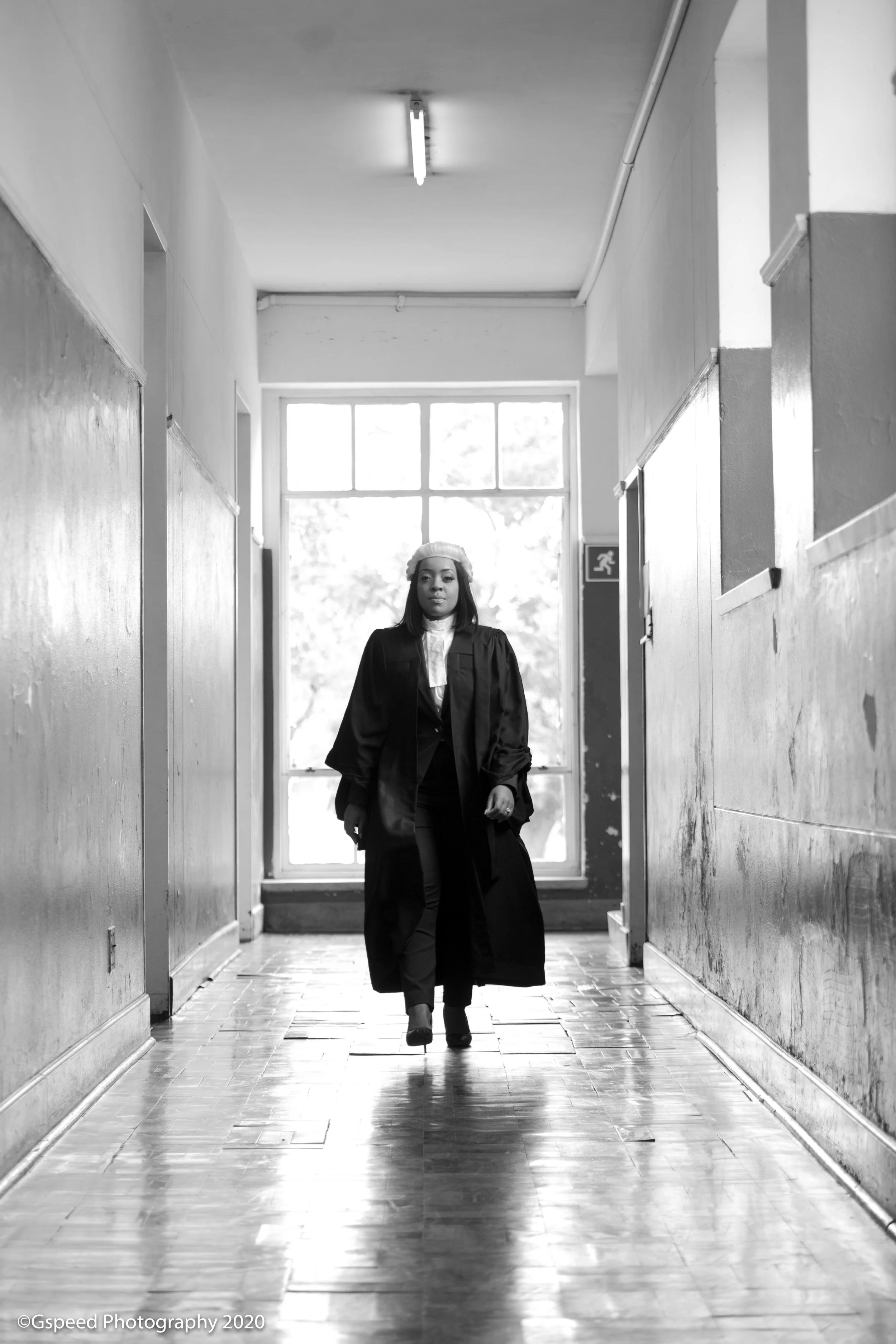 A woman in a graduation cap and gown walking down a hallway with wooden walls and a window at the end.