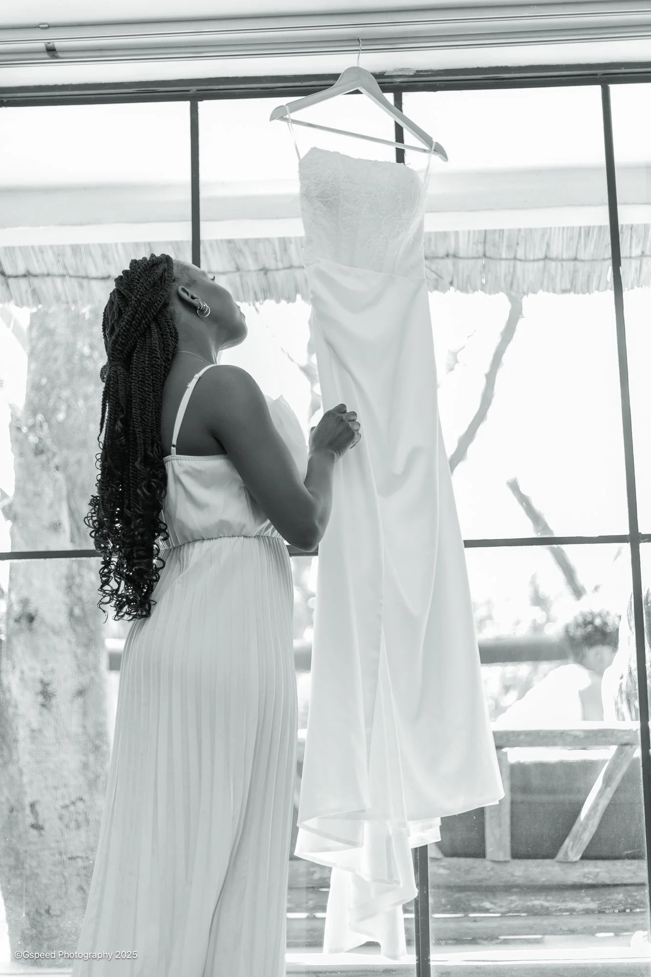 A woman with braided hair and earrings looking at a hanging wedding dress in front of a large window with trees outside.