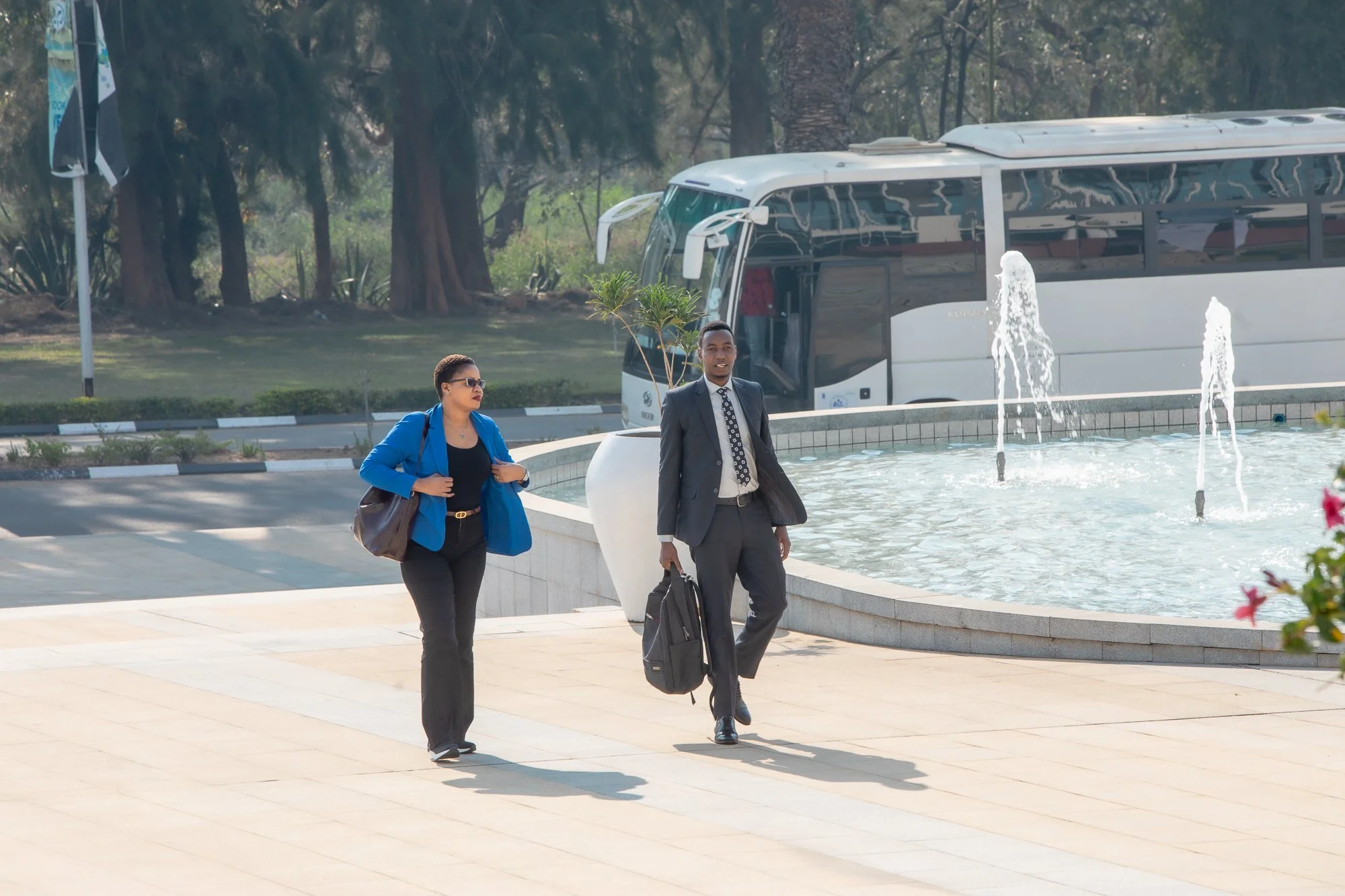 Two professionally dressed individuals, a woman and a man, walking past a fountain in an outdoor area with trees and a bus in the background. The woman is wearing sunglasses and a blue blazer, carrying a brown bag, and the man is wearing a suit and t