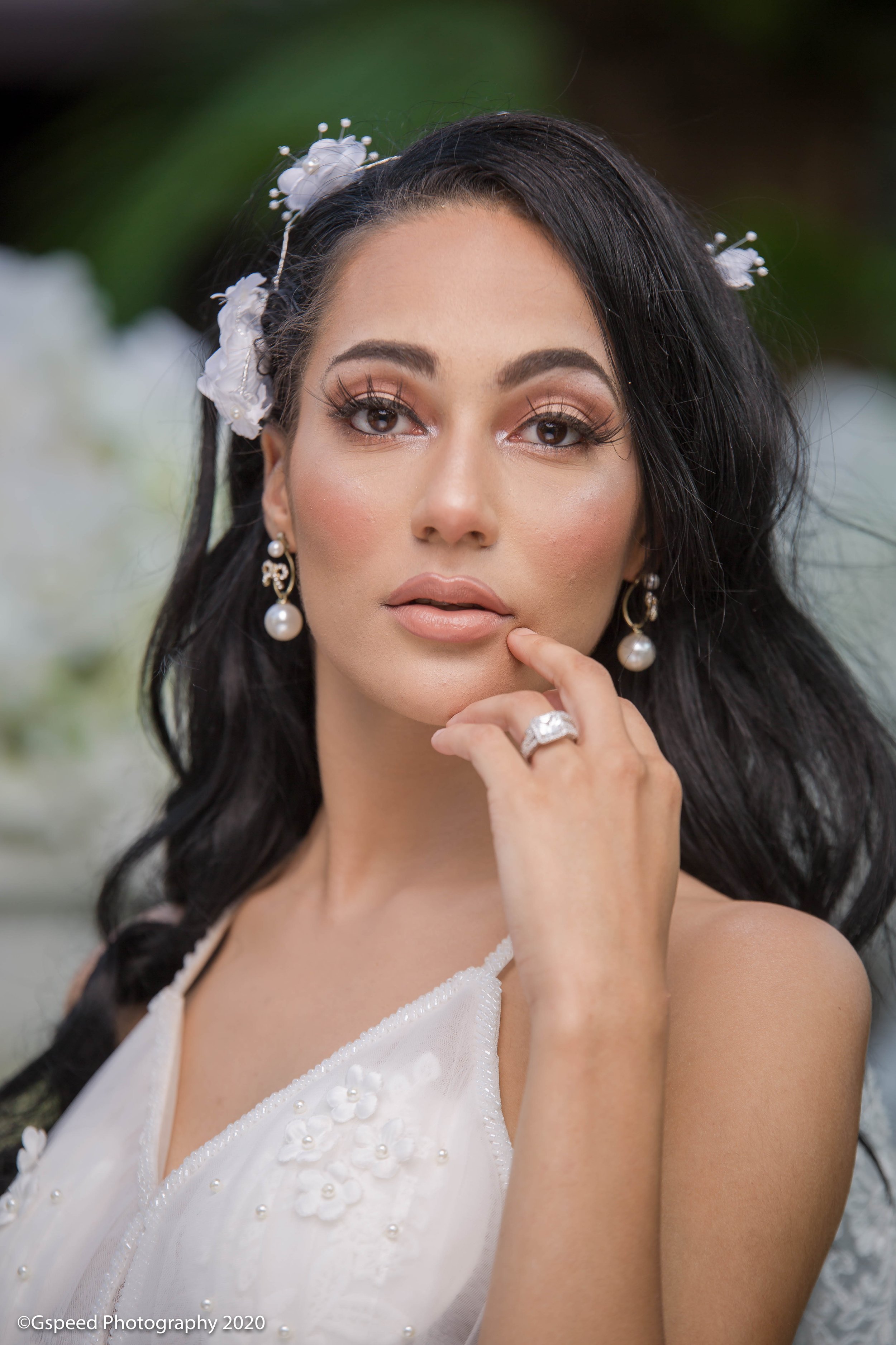 Close-up of a woman with dark hair wearing pearl earrings and a white dress, with decorative flowers in her hair.