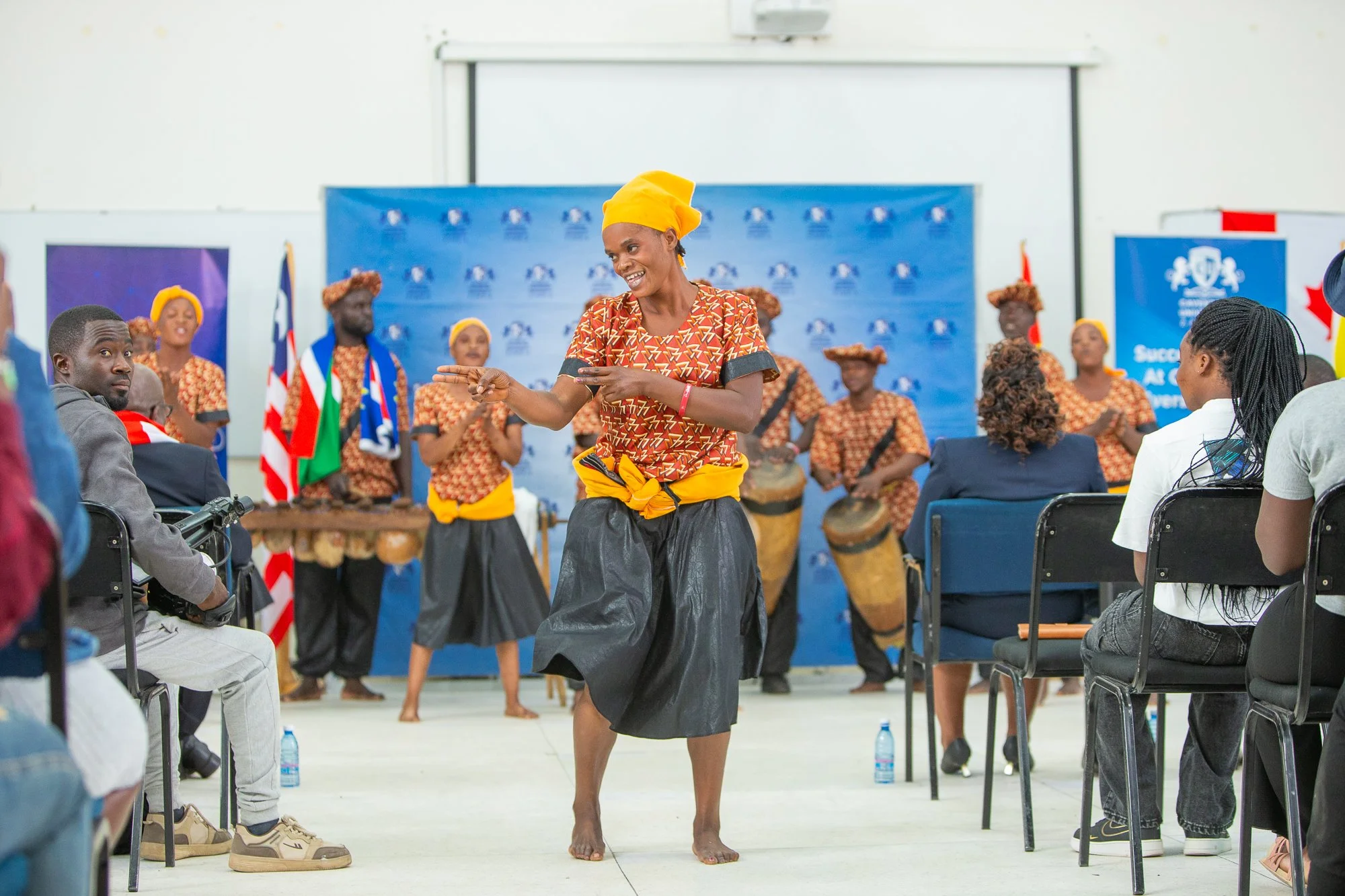 A woman dressed in traditional attire performs a dance in front of seated and standing audience members and drummers. In the background, there are flags and a blue backdrop with logos.