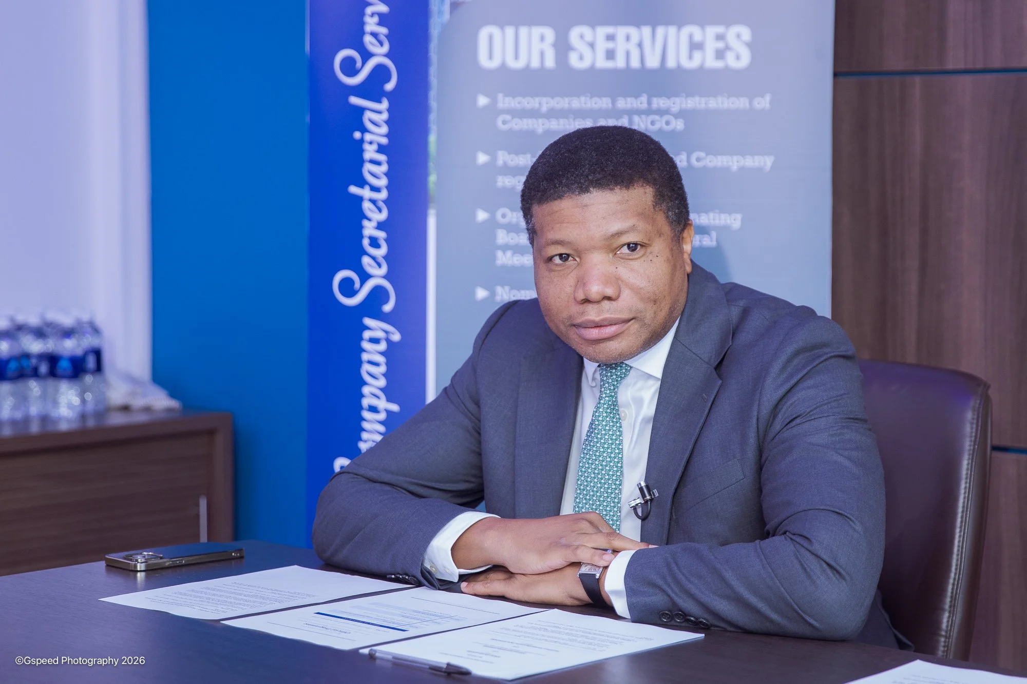 A man in a gray suit, white shirt, and green tie sitting at a desk in an office or conference room, facing the camera. Behind him is a blue banner with white text about services for incorporation and registration of companies and NGOs. There are pape