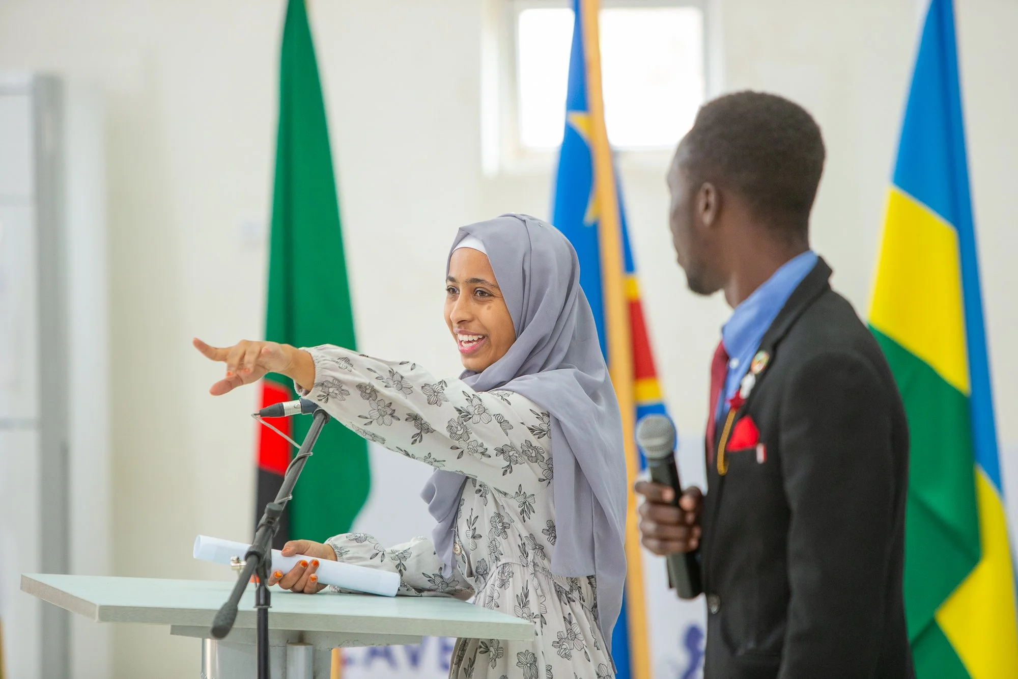A woman wearing a gray hijab and floral dress speaking at a podium with a microphone, gesturing with her hand, during a formal event with flags in the background. A man in a suit holding a microphone stands nearby.