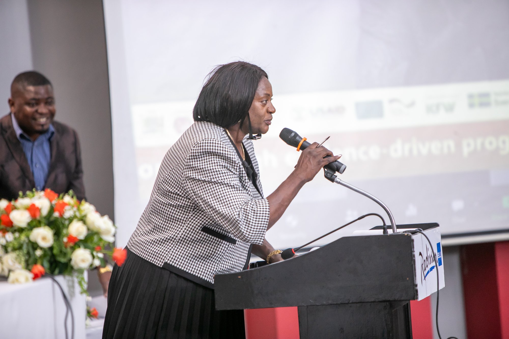 A woman speaking into a microphone at a lectern during a conference or event, with a man smiling in the background and a large screen behind them displaying a presentation slide.