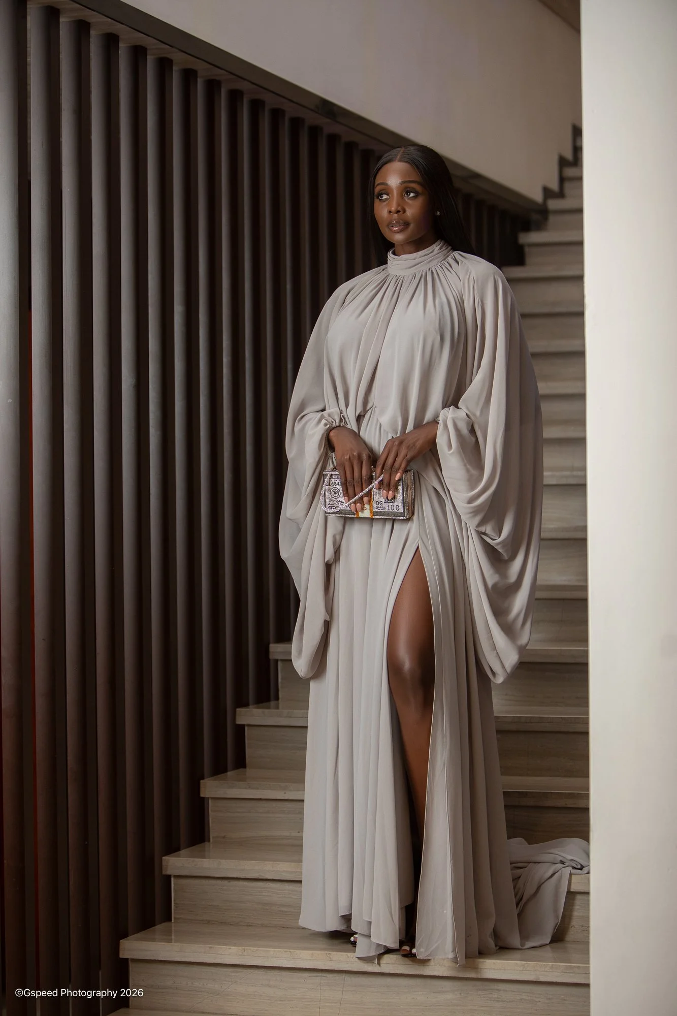 A woman in an elegant beige gown with a high slit, holding a glittery clutch bag, standing on wooden stairs near a wall with dark vertical slats.