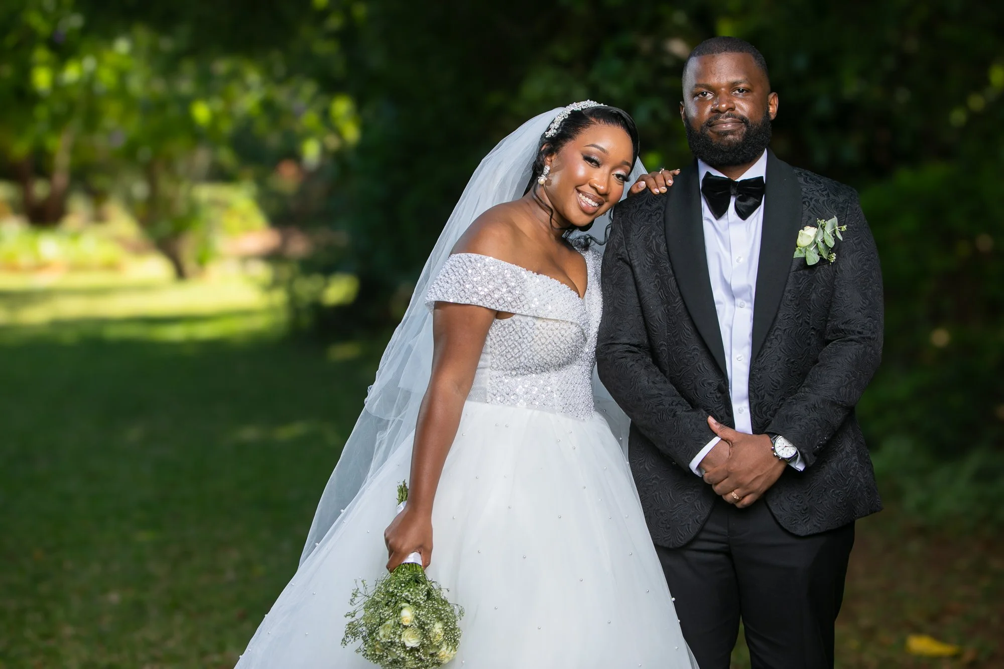 A bride and groom posing outdoors on a sunny day, with the bride wearing a white wedding dress and veil, holding a small bouquet of white roses and greenery, and the groom in a black tuxedo with a bow tie and boutonniere, standing close and smiling.