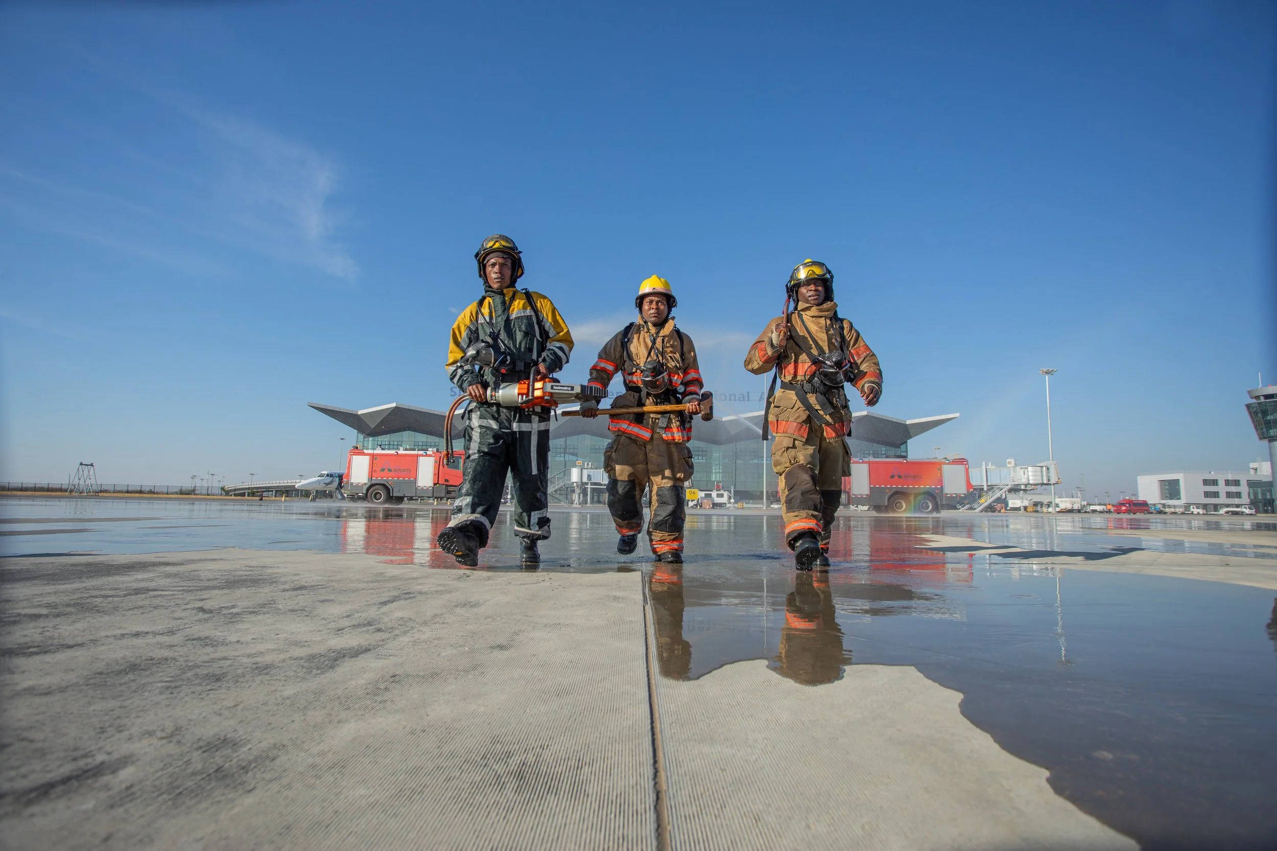 Three firefighters walking on the airport tarmac with a reflection visible on a wet surface, buildings and a support vehicle in the background under a clear blue sky.