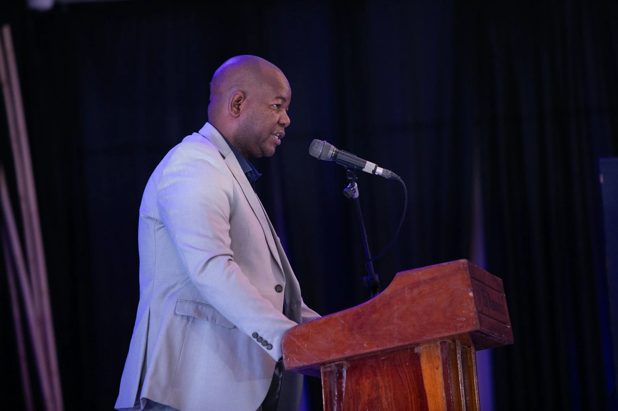 A man in a light-colored suit jacket is speaking at a podium with a microphone in front of a dark background.