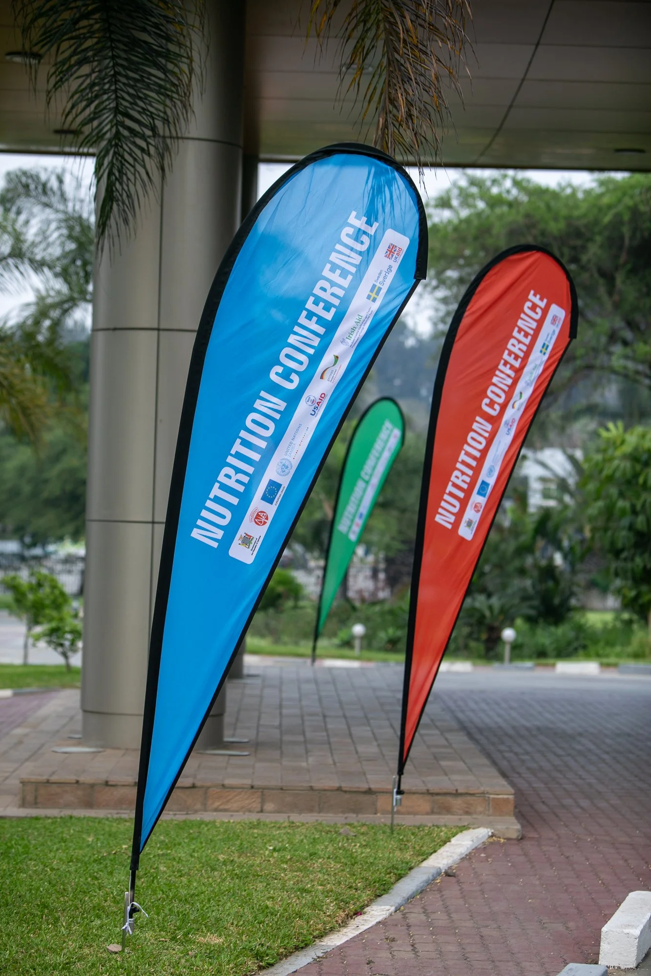 Colorful flags with registration for a nutrition conference, including logos for the European Union, Ireland, Sweden, and other sponsors, set outdoors on a city sidewalk with trees and a building in the background.
