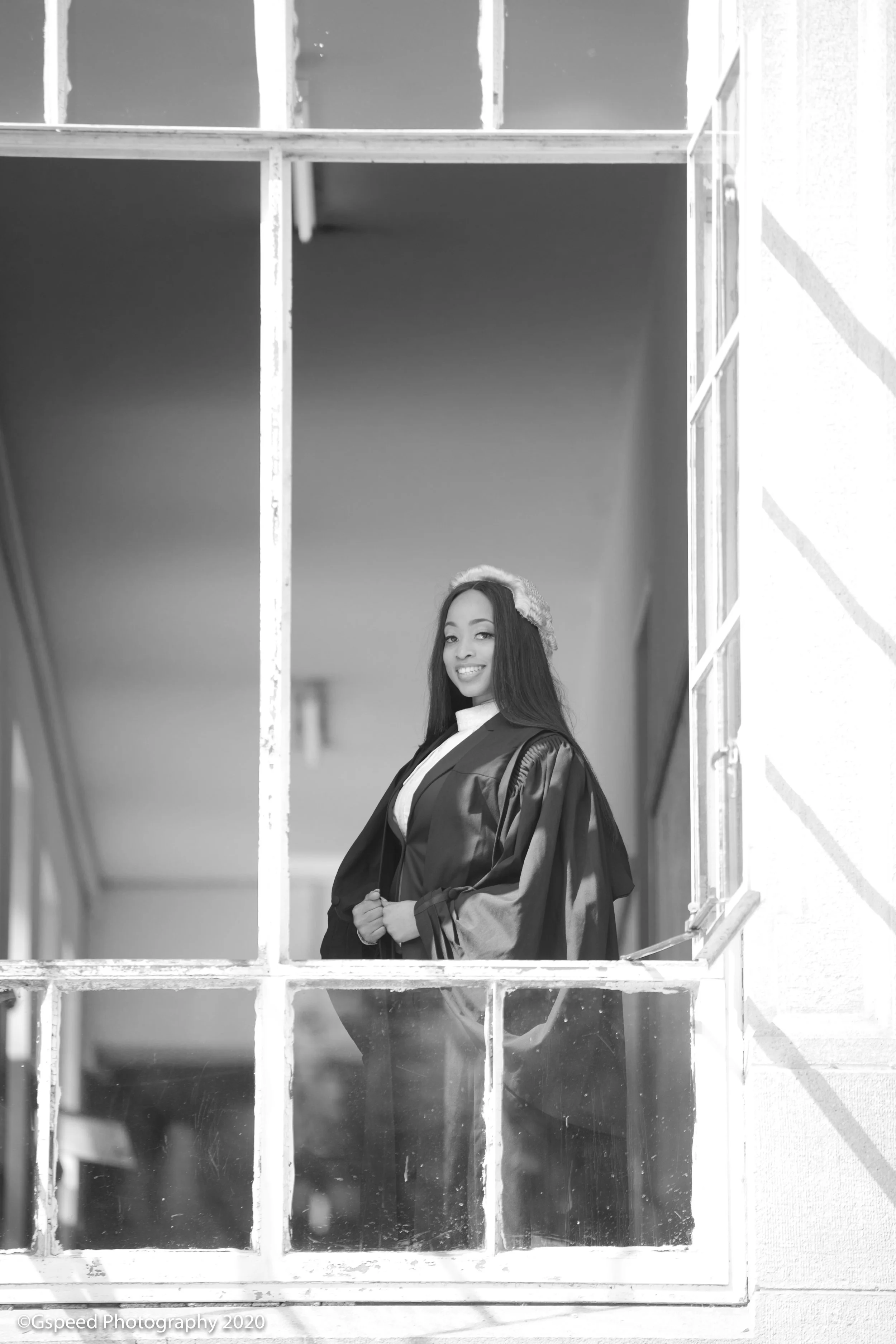 Black and white photo of a young woman in graduation gown and cap, standing behind a window with divided panes, smiling at the camera.