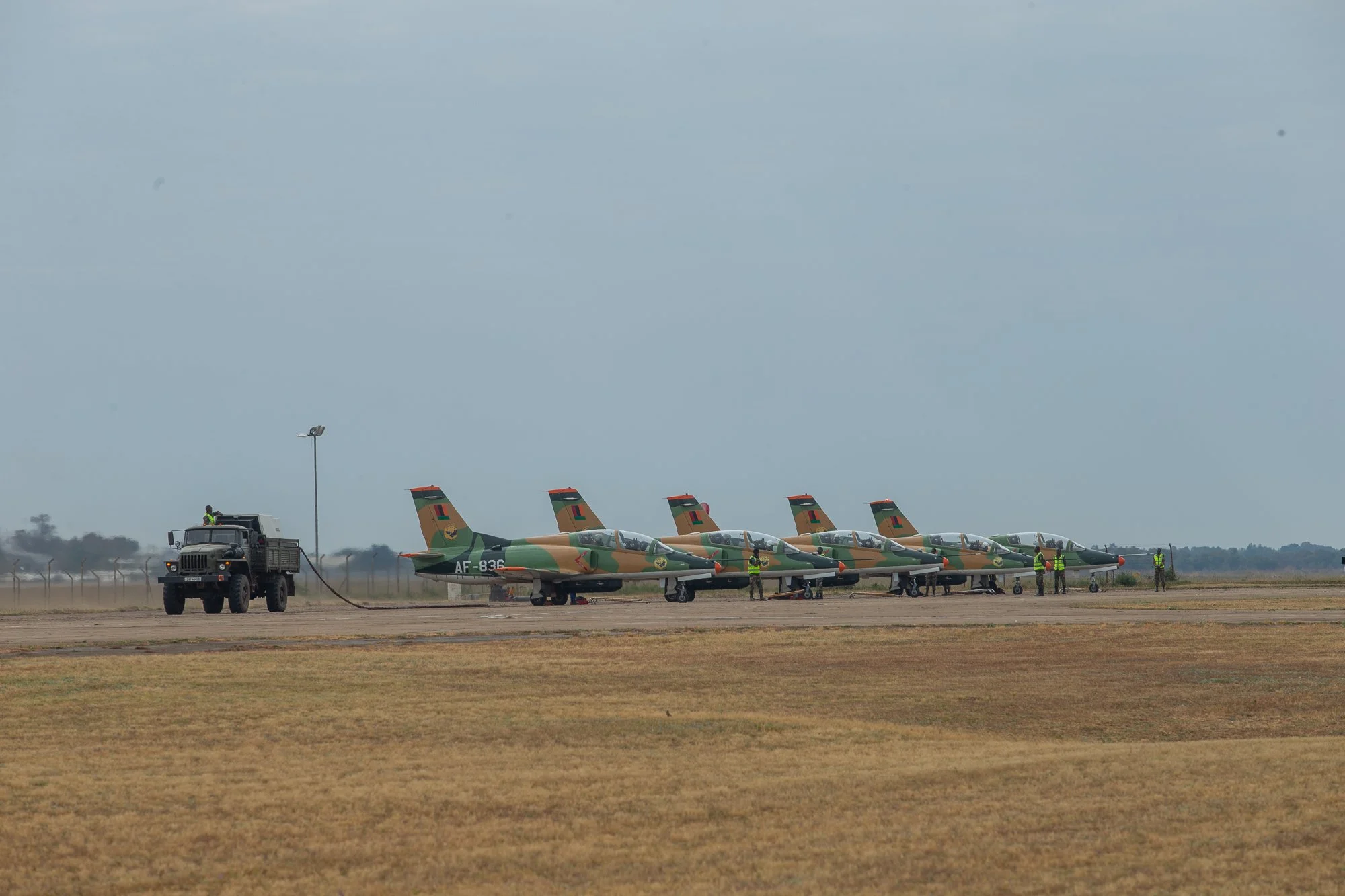 Military jets painted in camouflage colors parked on runway with personnel nearby, a truck connected to one of the jets, and a cloudy sky overhead.