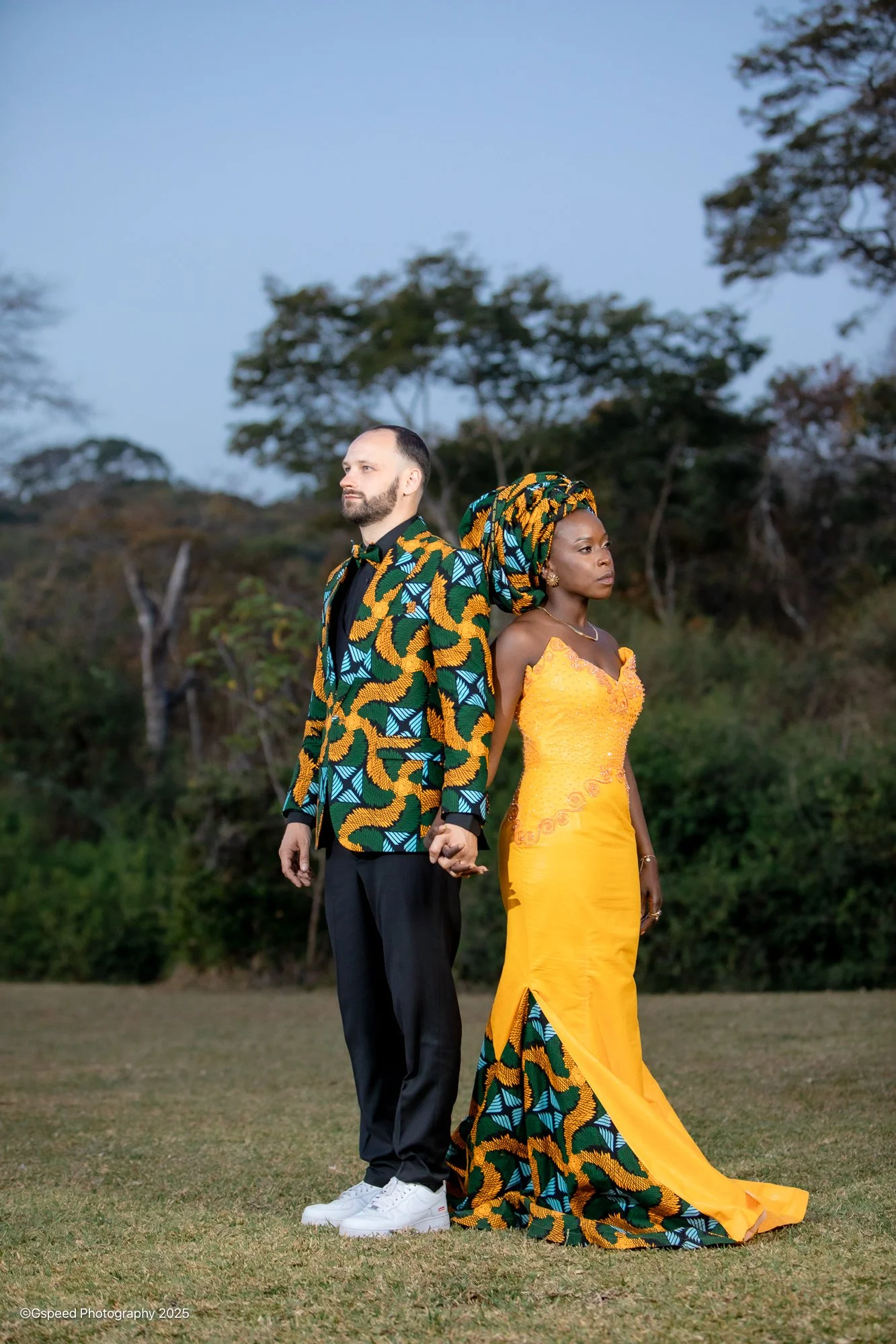 A man and woman standing outdoors on a grassy field with trees in the background. The man is wearing a colorful patterned suit with a black shirt and white sneakers, and the woman is dressed in a bright yellow dress with matching traditional headwrap