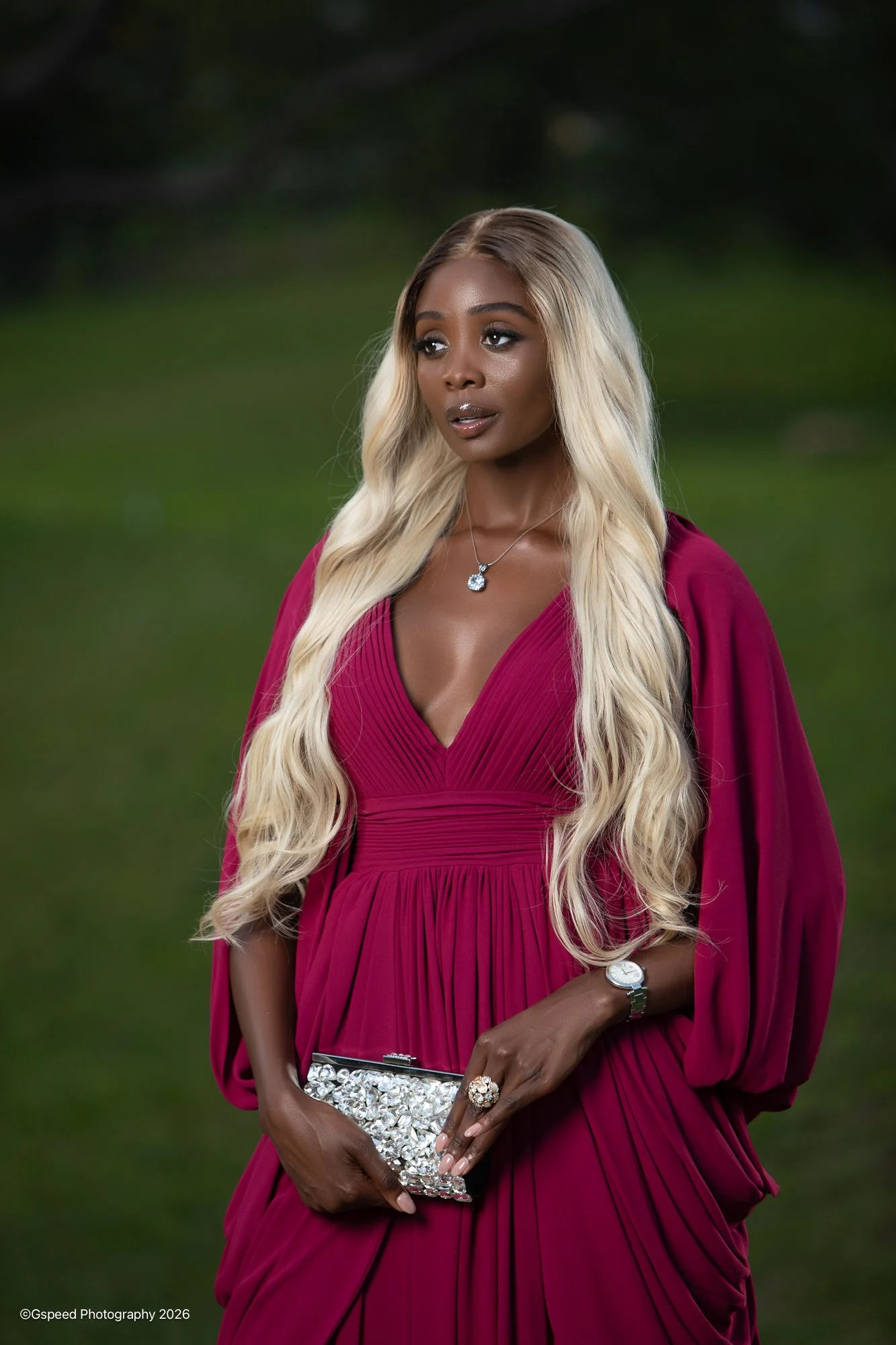 A woman with long blonde hair wearing a deep red dress with pleats and puffed sleeves holding a bejeweled clutch bag and wearing a necklace, standing outdoors on a grassy area.