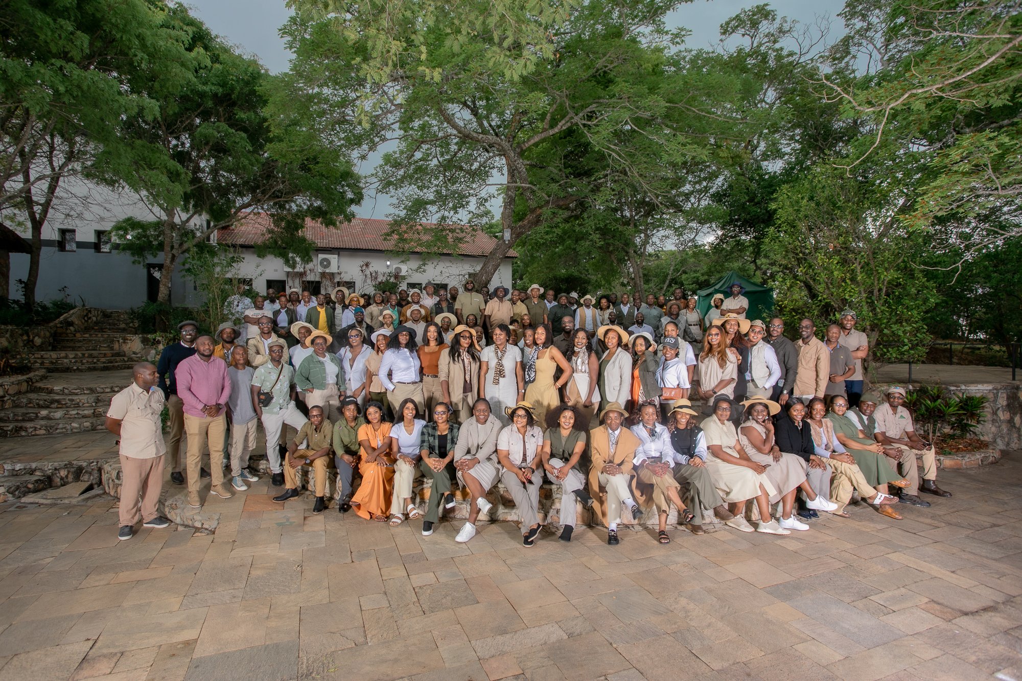 A large group of people posing for a photo outdoors in front of trees and a white building with a red tile roof. The group includes men and women of various ages, many wearing casual clothing and hats, with some seated in front and others standing be