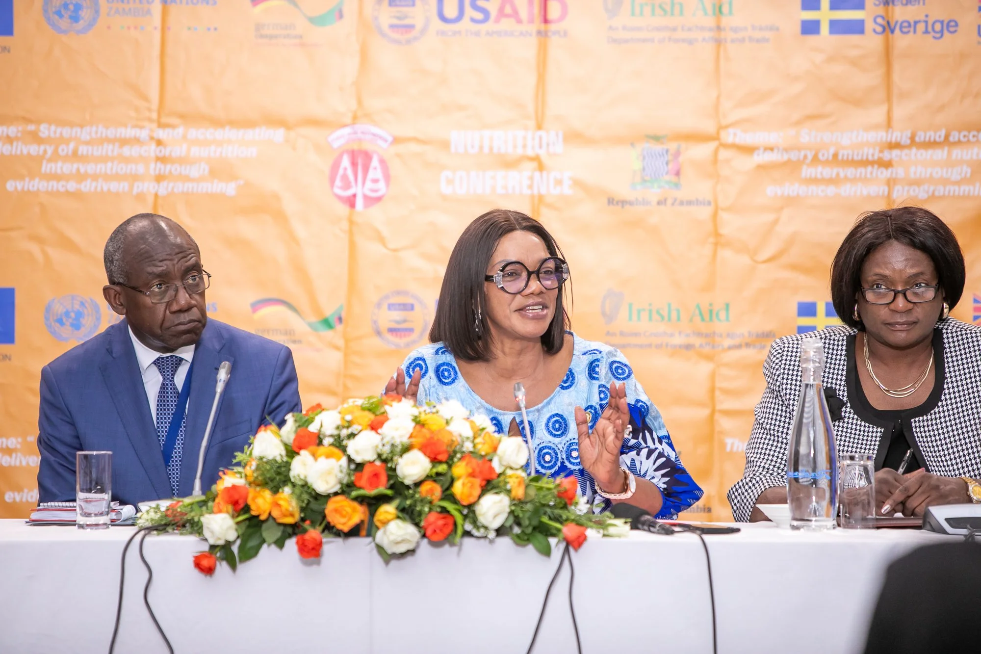 Three people sitting at a conference table with a large flower arrangement in front of them. A man in a blue suit and glasses on the left, a woman in a blue and white patterned dress with glasses in the center, and a woman in a black and white checke