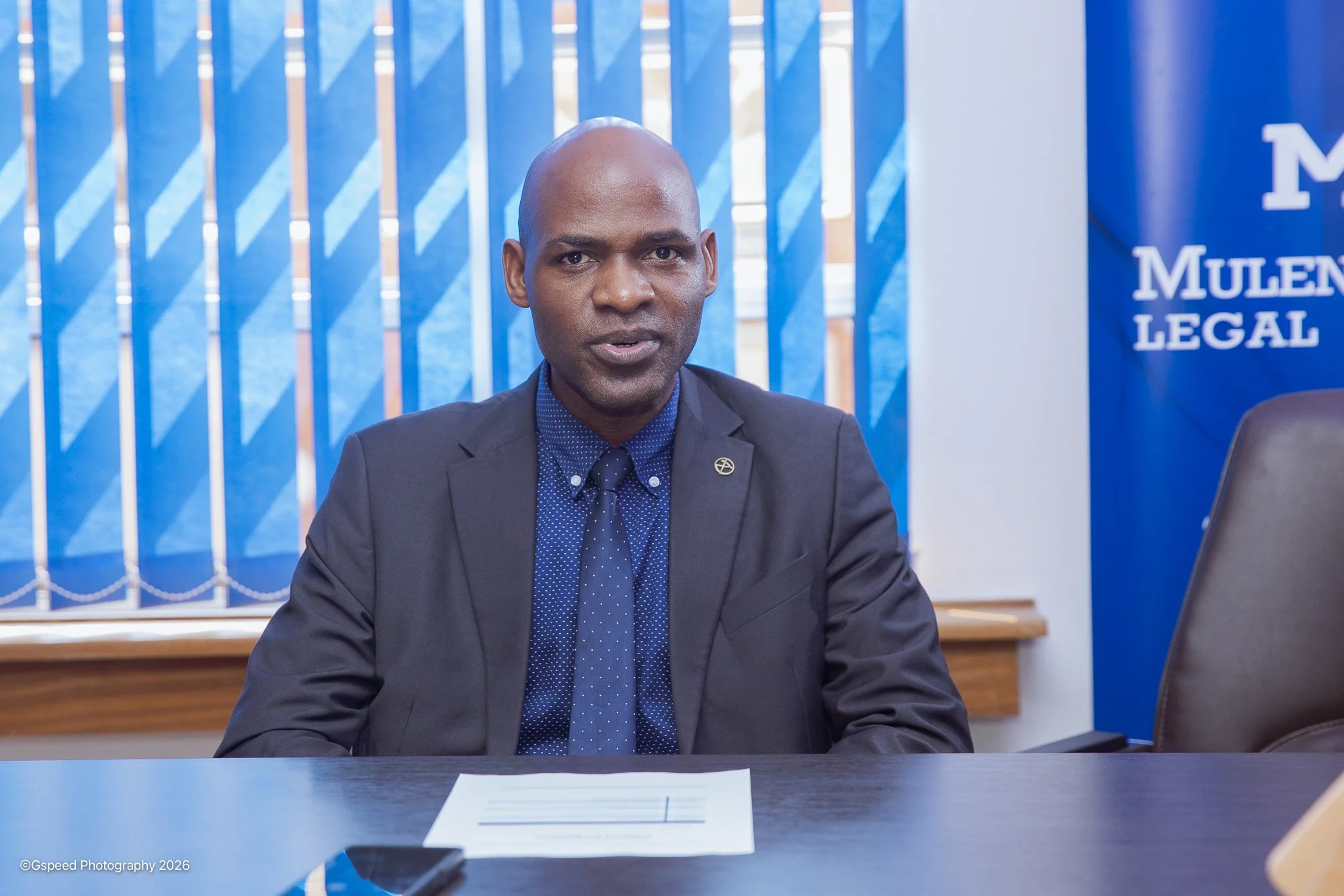A man in a business suit sitting at a desk during a meeting with a conference room background, including a blue banner with the words "MULEN LEGAL" and a blue backdrop with vertical blinds.