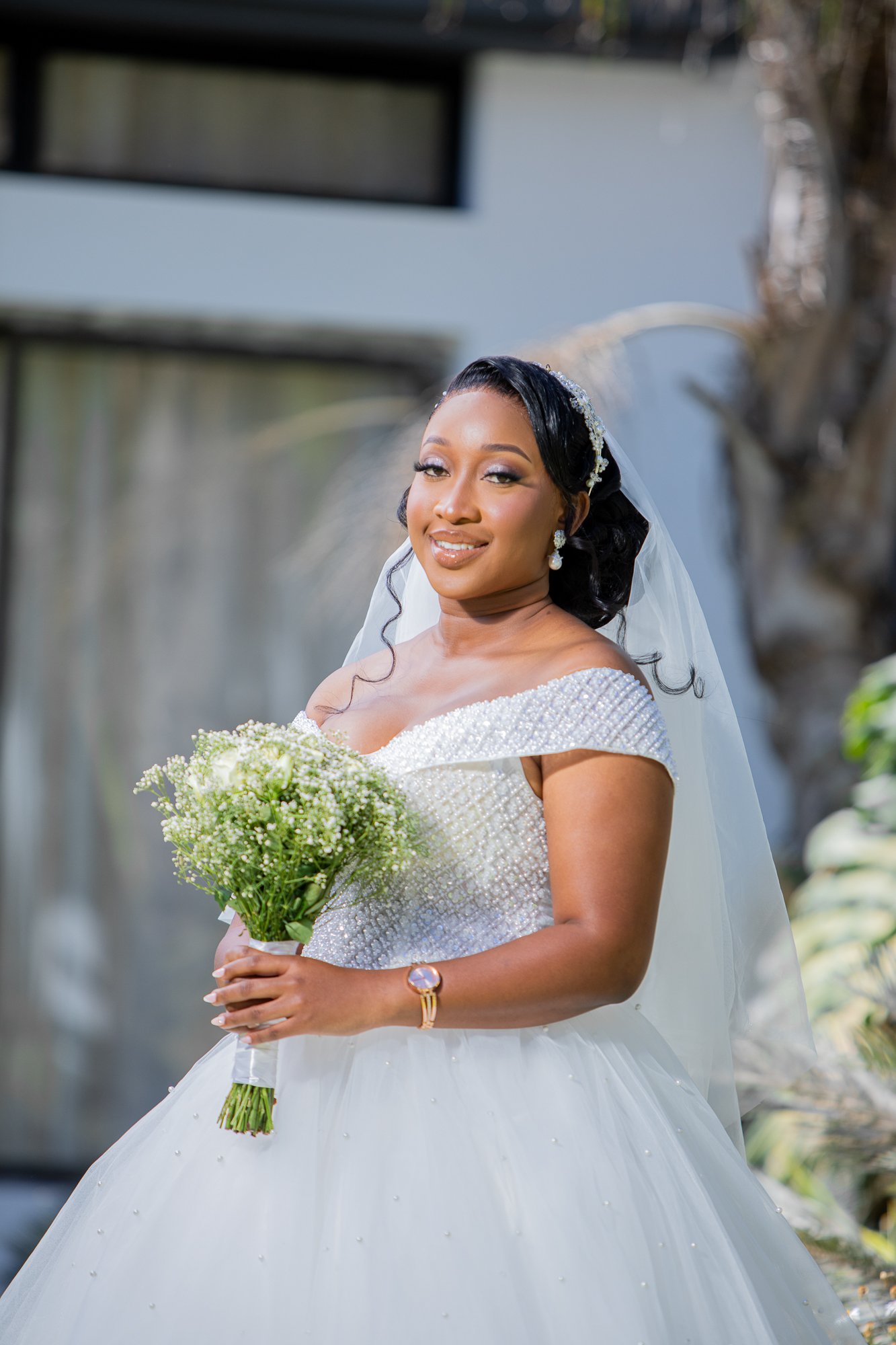 A bride in a wedding dress holding a bouquet of flowers outdoors, smiling and looking slightly to the side.