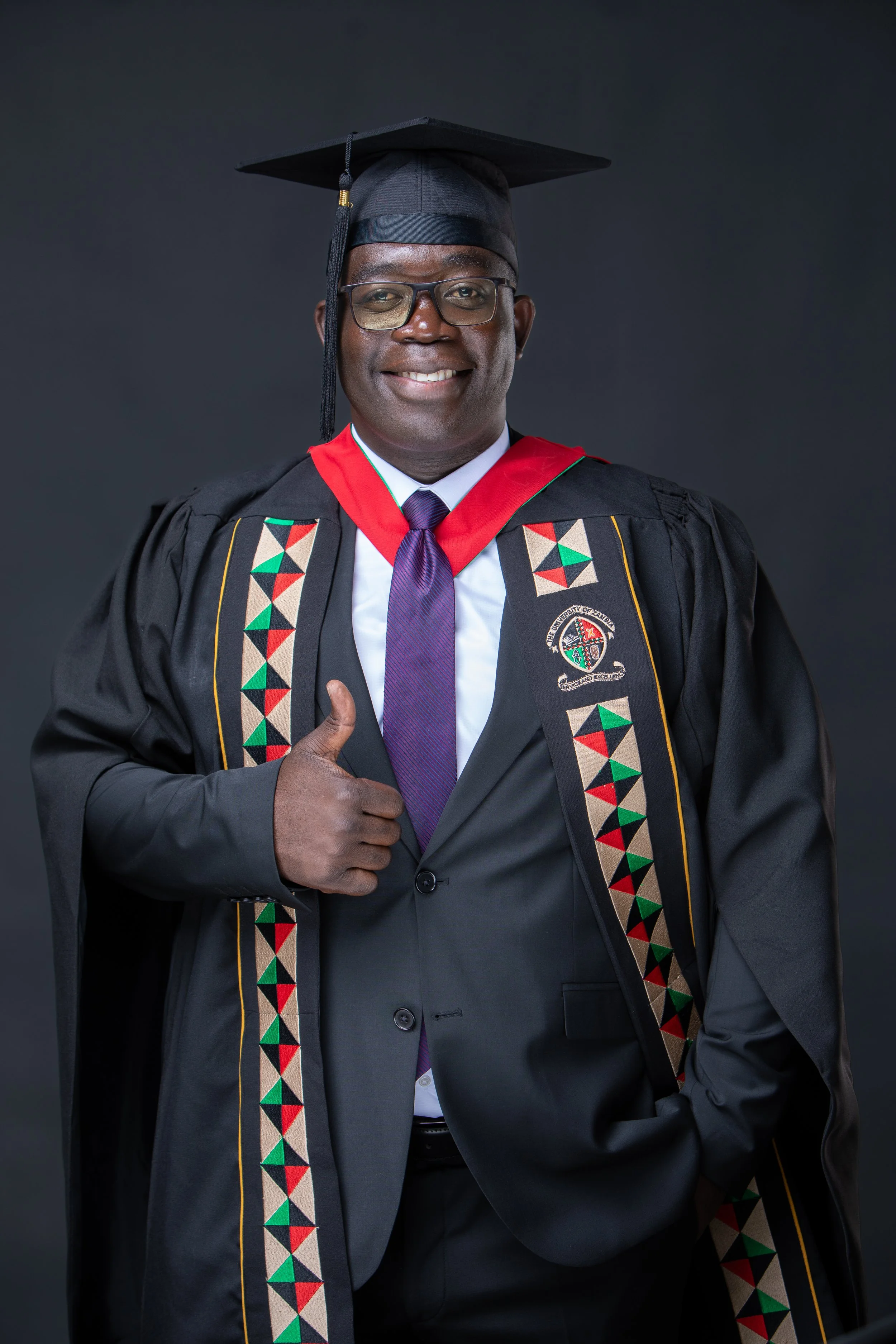 Portrait of a man in graduation attire, wearing a black gown with colorful traditional African patterns, a black cap, glasses, a purple tie, and a white shirt, standing against a dark background, smiling and giving a thumbs up.