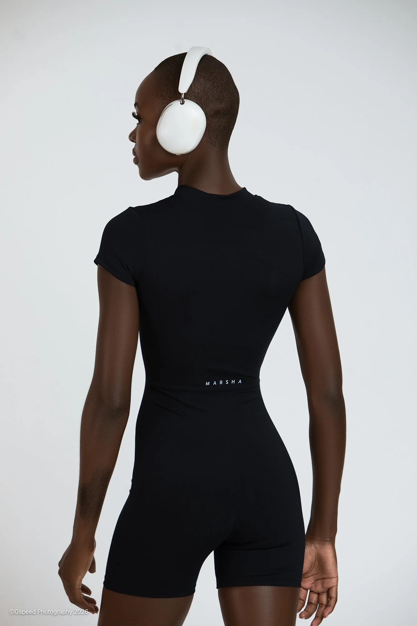 A woman with dark skin and a shaved head is seen from behind wearing a black outfit and white headphones, standing against a plain light background.