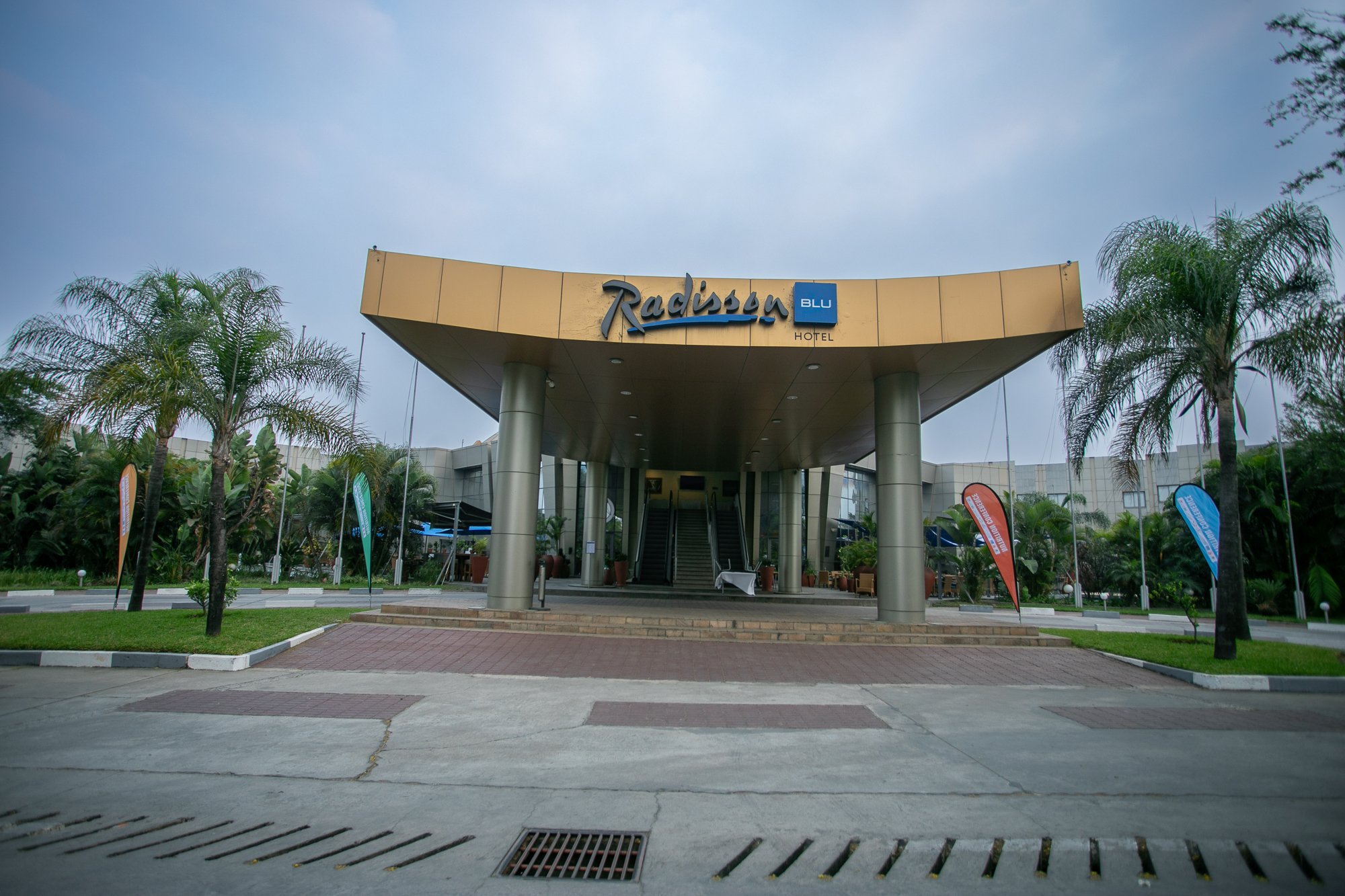 Entrance of Radisson Blu hotel with a canopy, palm trees, and flags, under a cloudy sky.
