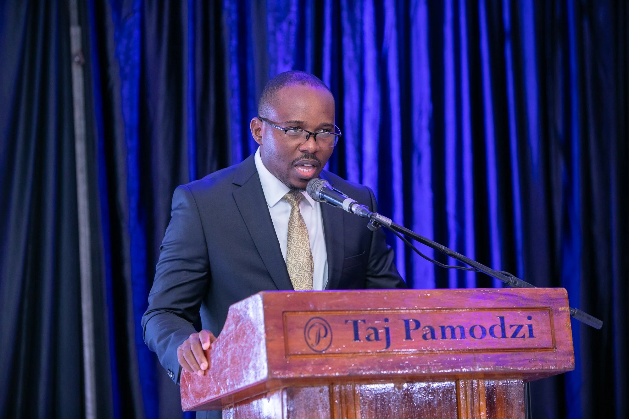 Man in business suit and glasses speaking at a podium with a microphone, dark curtain background.