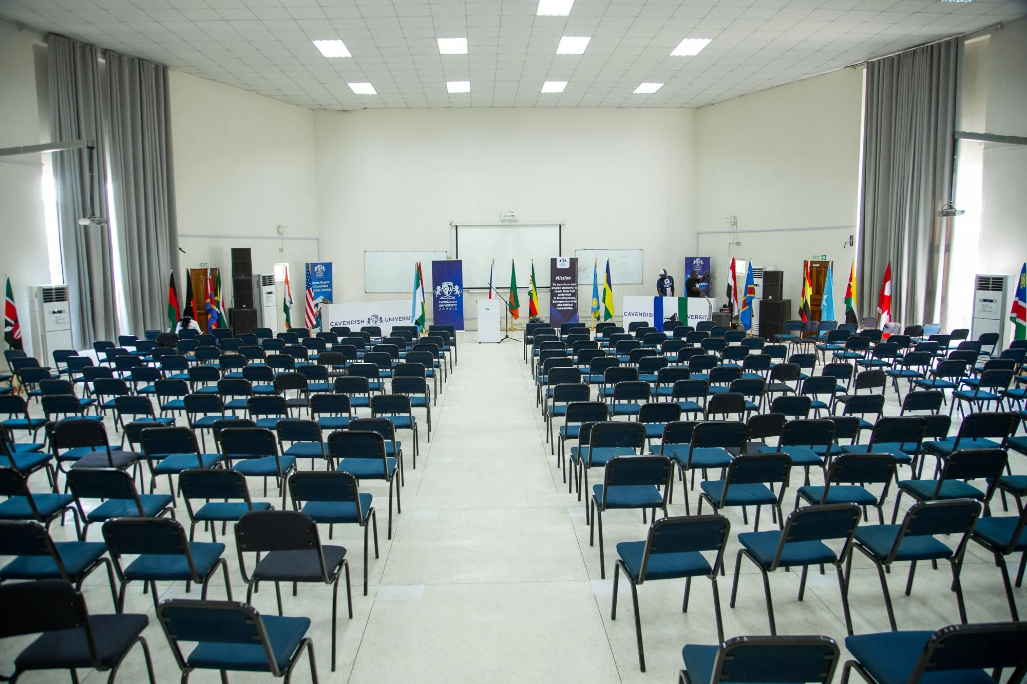 Large auditorium or conference hall with rows of black chairs facing a stage with flags and banners. The stage has a podium, whiteboards, and two individuals preparing equipment. The room has white walls, large windows with curtains, and air conditio