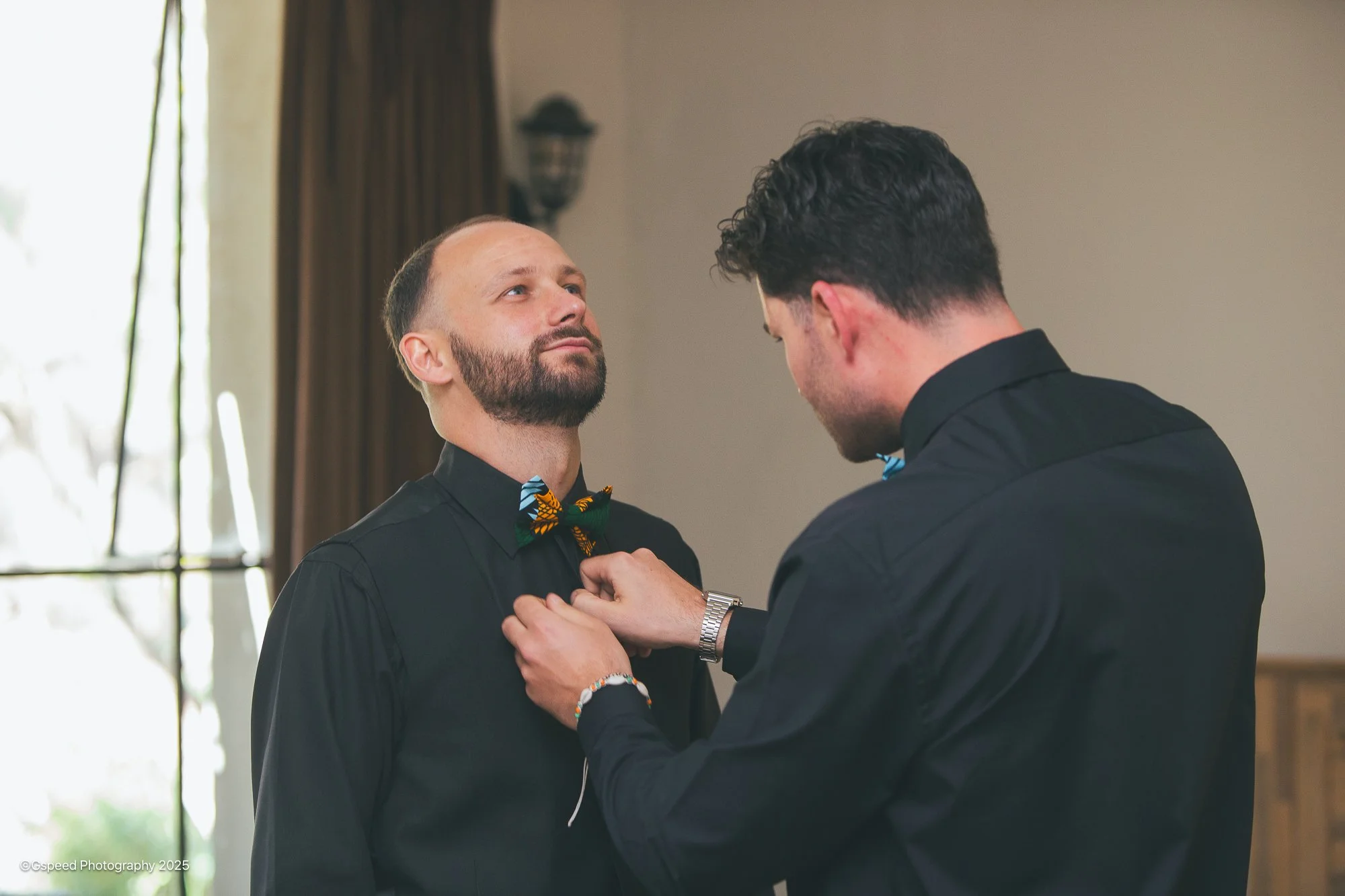 Two men in black shirts and bow ties, one with a beard and the other with curly hair, during a wedding ceremony or event, with one man pinning a boutonniere on the other's shirt.