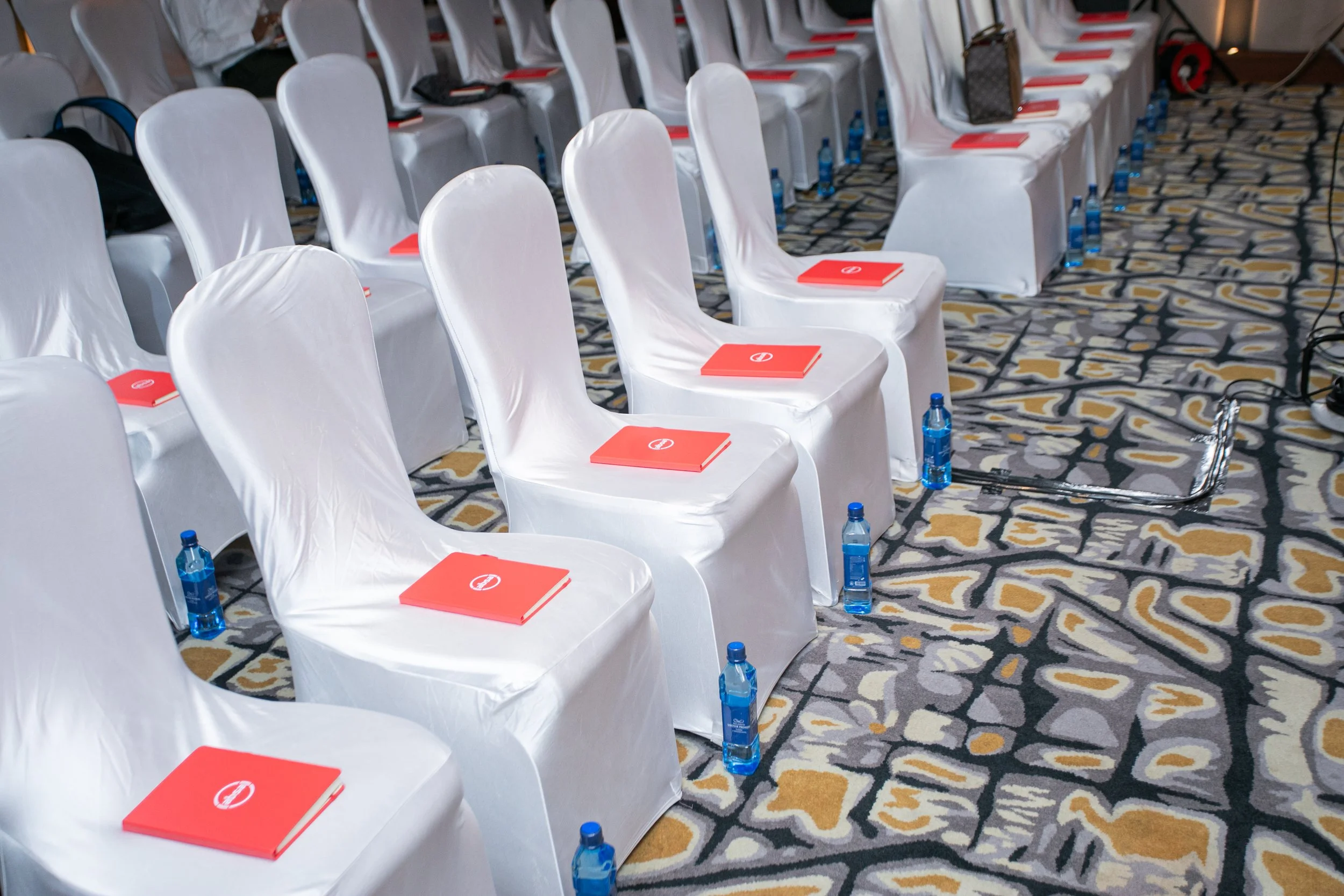 Rows of white chairs covered with white cloths, each with a red booklet and a blue water bottle placed beside them, set up in a conference or meeting room with a patterned carpet.