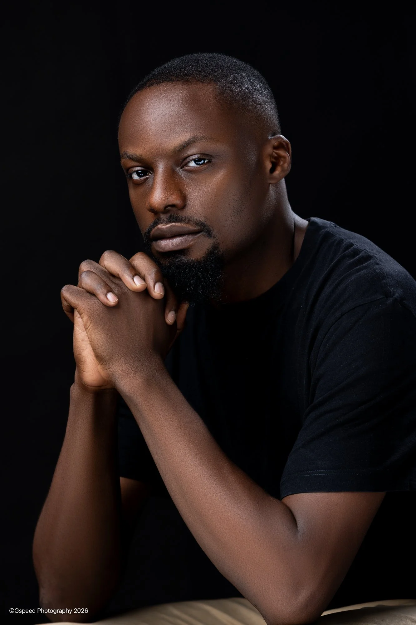 A young Black man with short hair and a beard, wearing a black shirt, posing against a black background with his hands clasped near his face.