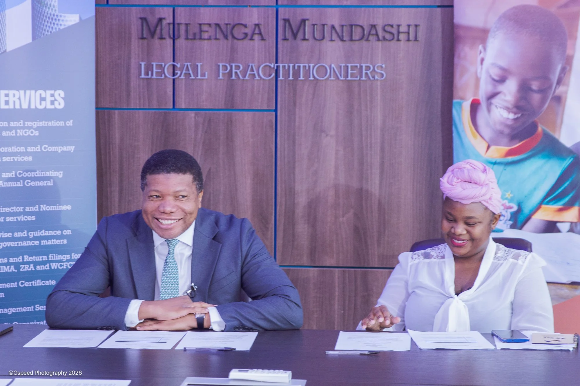 Two people sitting at a desk with documents, a pen, and a calculator, smiling. Behind them, there is a large sign reading 'Mulenga Mundashi Legal Practitioners' and a woman on a poster smiling.