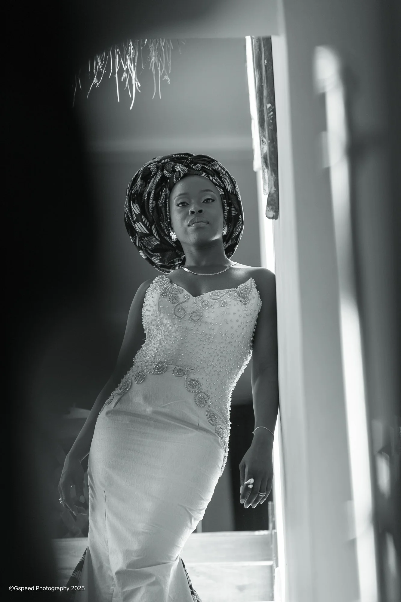 Black and white photo of a woman in a detailed wedding dress and patterned headwrap, standing at a window with light shining through, looking confidently at the camera.