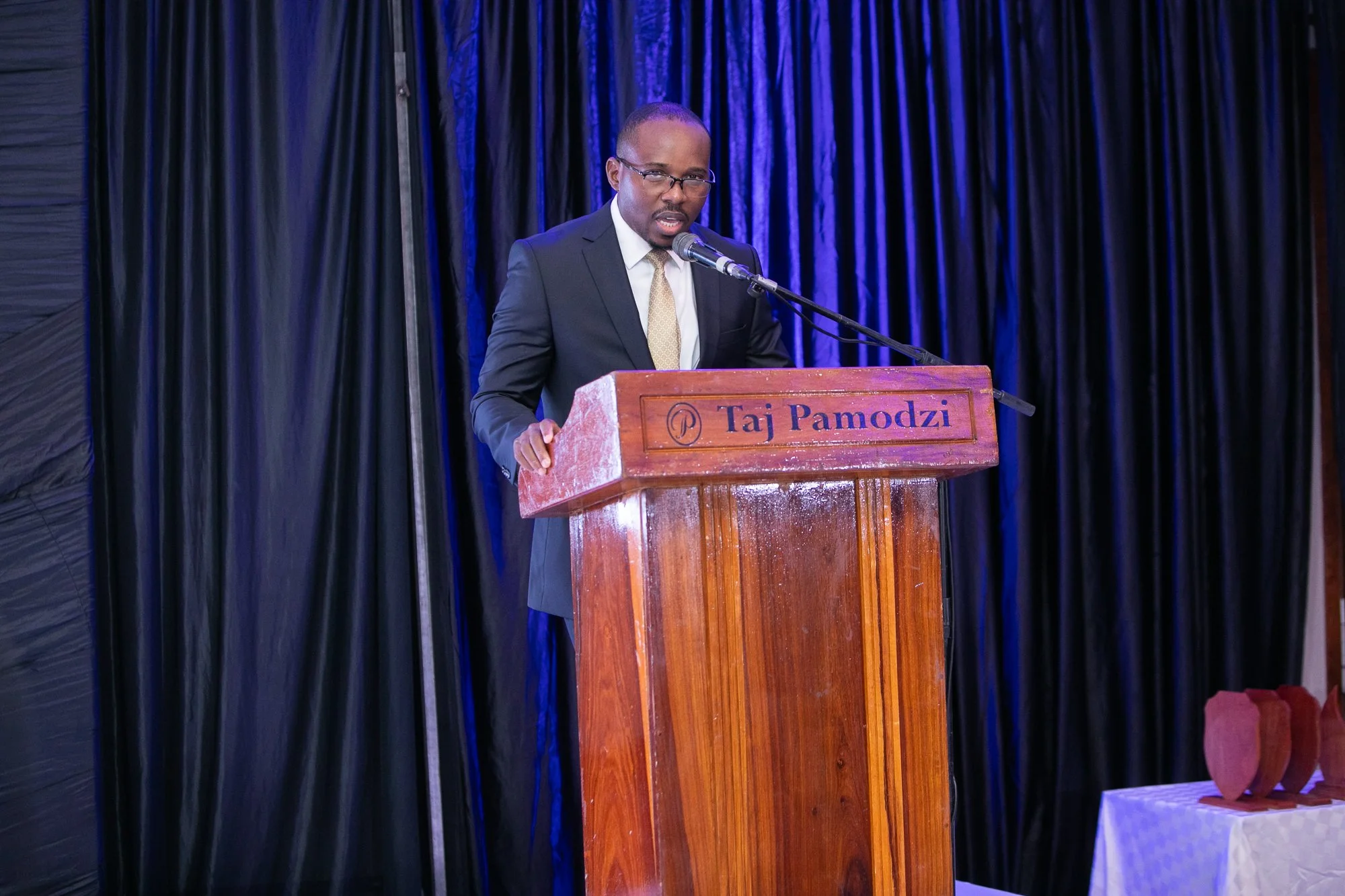 A man in a suit and glasses speaking at a podium with a microphone, with a dark curtain backdrop, at an event named Taj Pamodzi.