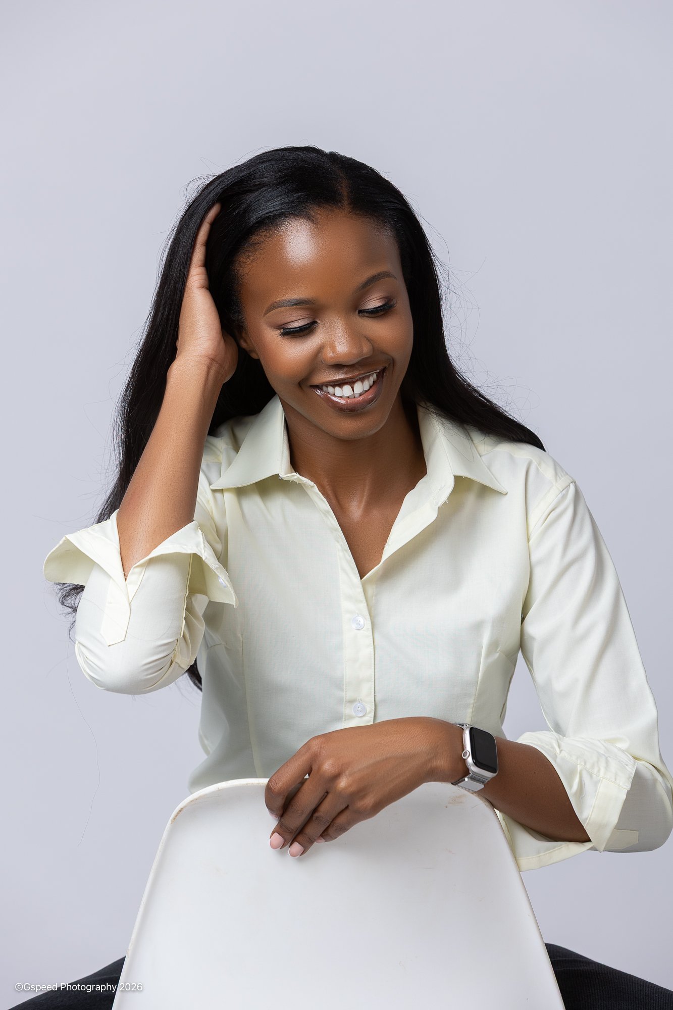 A woman with long black hair, wearing a light yellow button-up shirt, is sitting on a white chair, smiling with eyes closed, touching her hair with her left hand and resting her right arm on the chair. She also wears a smartwatch on her right wrist a