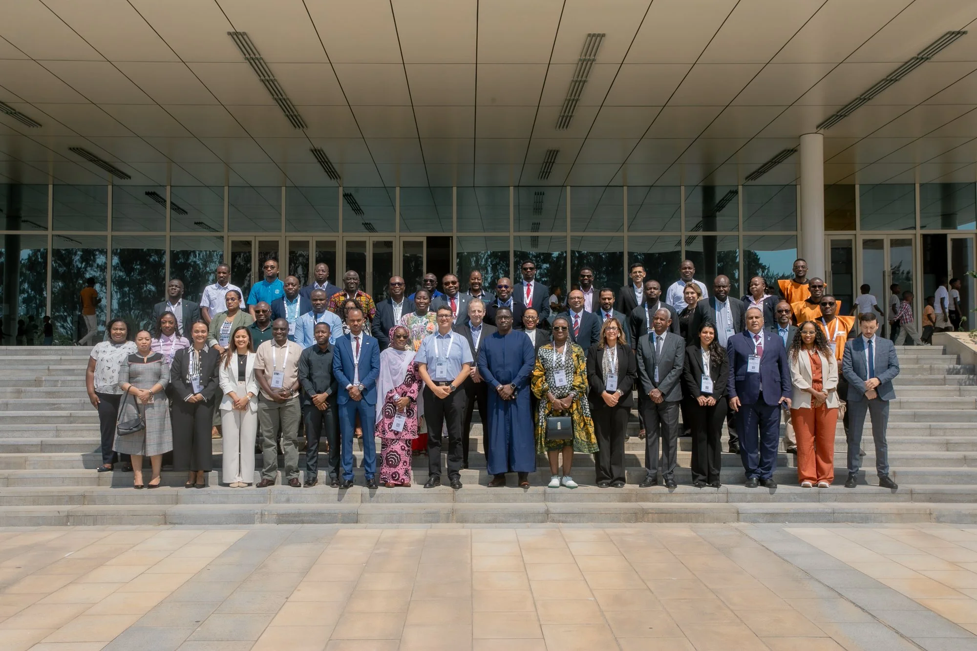 A large group of diverse men and women in formal and traditional attire standing on steps in front of a modern building with glass windows and a flat roof, posing for a group photo outside.