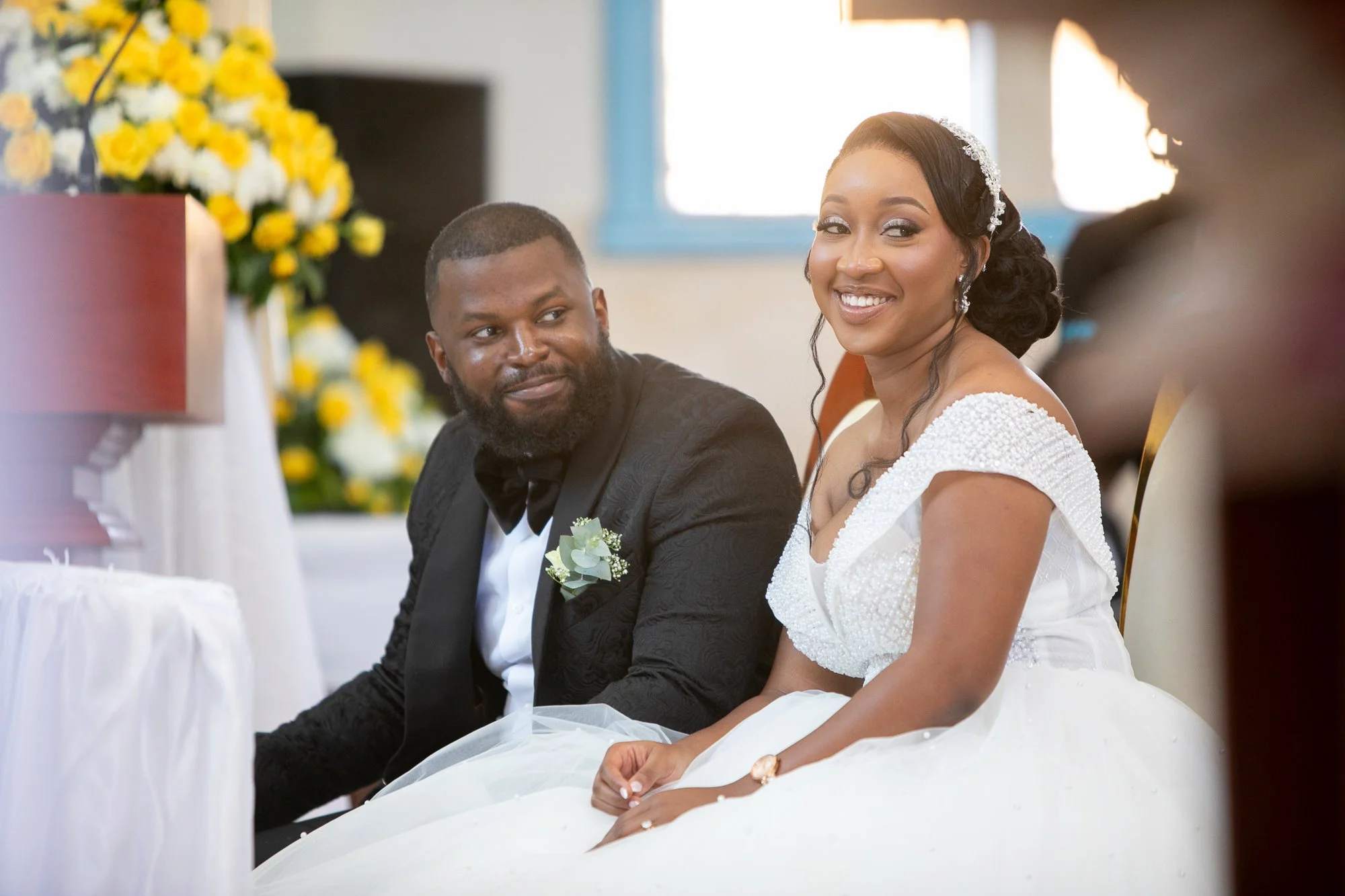 Bride and groom sitting at the wedding reception table, smiling and looking to the side, with a backdrop of yellow flowers and window light.