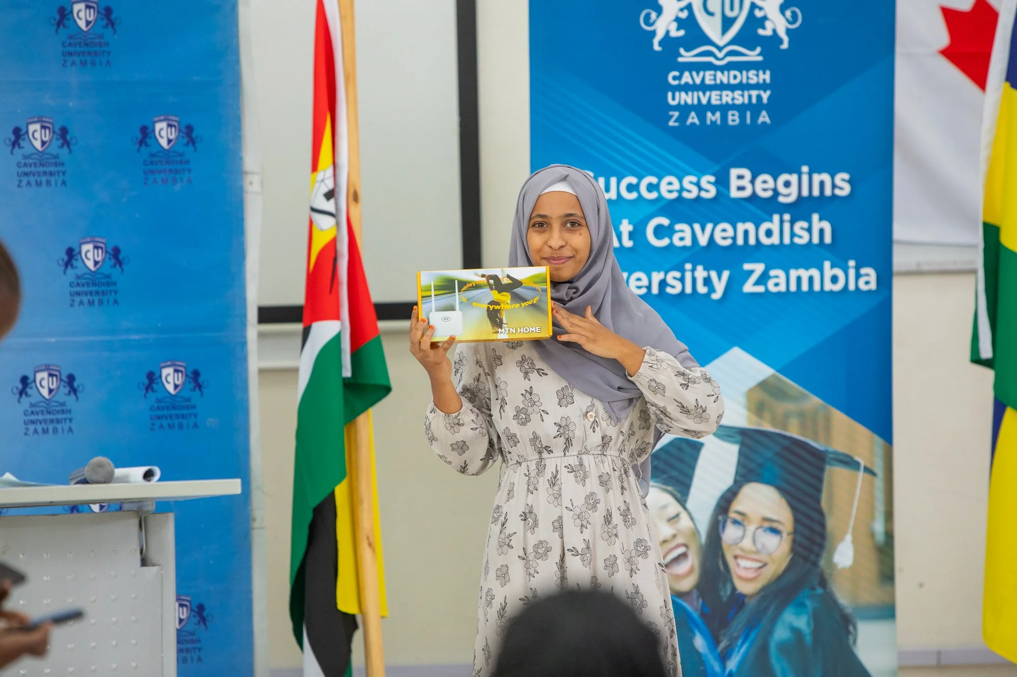 A young woman with a hijab holding a package labeled 'MTN Home' at a seminar or event at Cavendish University Zambia, with a blue backdrop and flags.