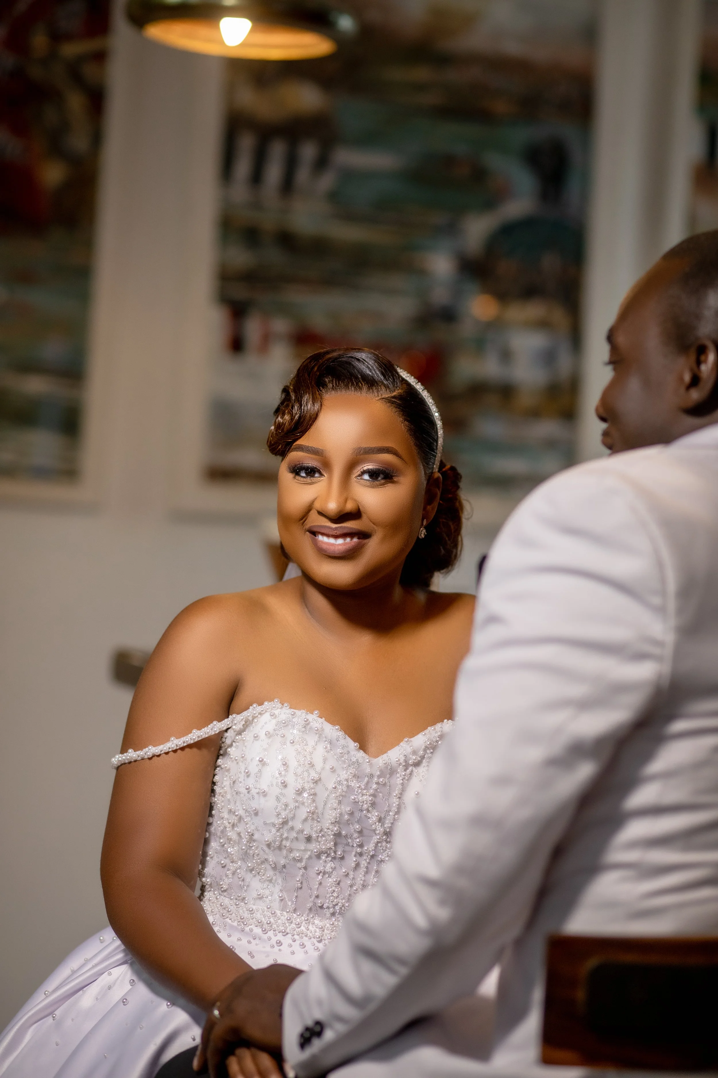 A smiling bride in a white wedding gown with bead embellishments, sitting and looking at her partner, a groom in a white suit, during a wedding ceremony inside a room with abstract artwork on the wall.